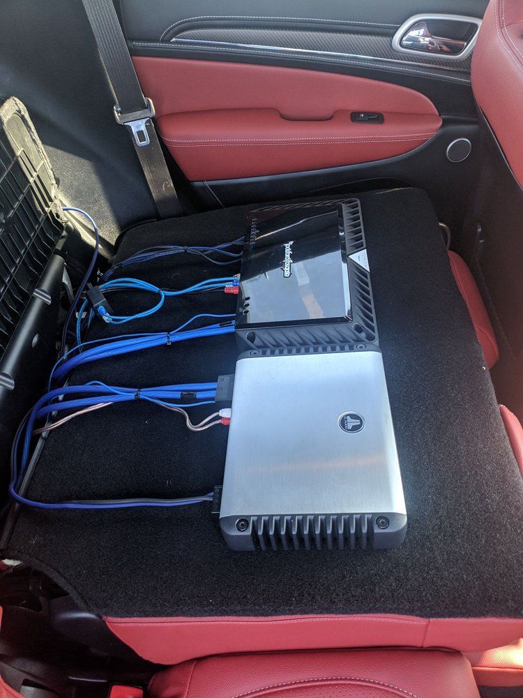 A car's back seat with installed audio equipment. Silver amplifiers sit on a black mat with various wires. The seat is red.