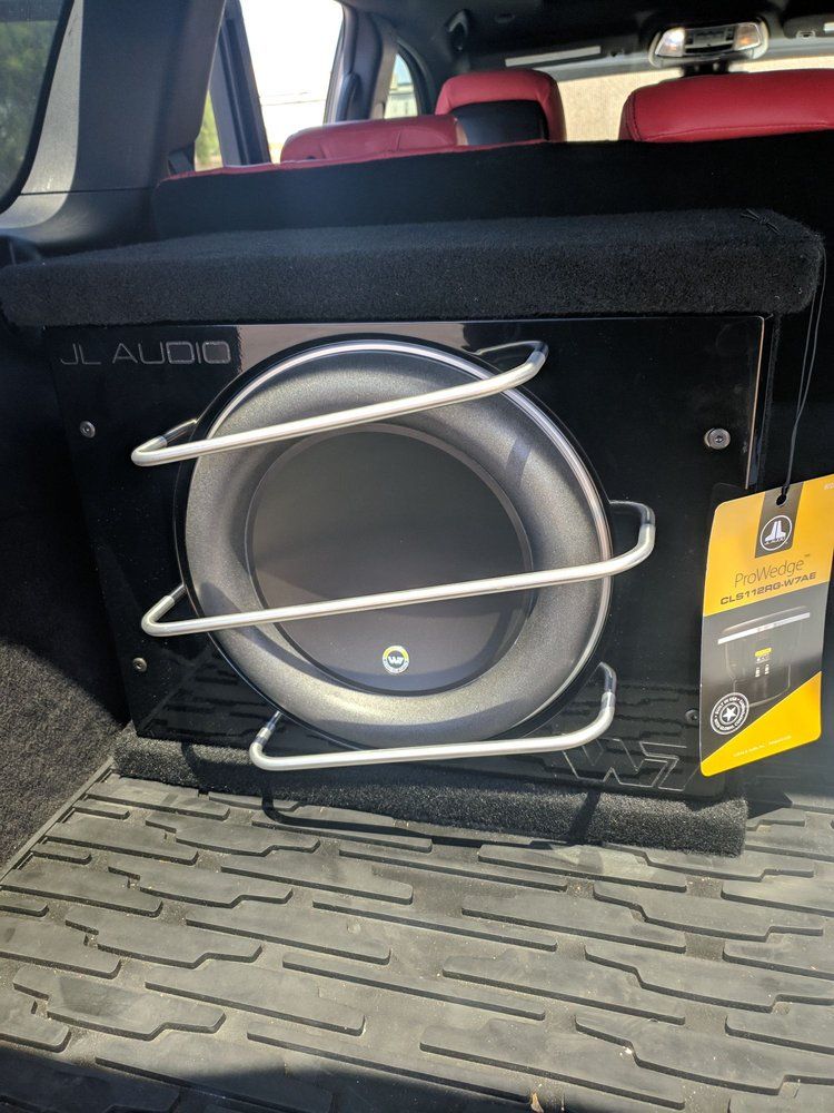 Black JL Audio subwoofer in the trunk of a car. It has a silver protective cage and a new tag.