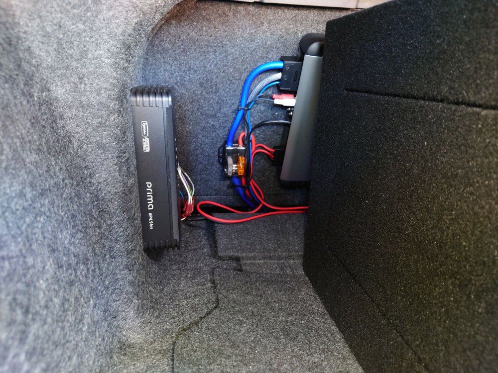Car trunk with a black amplifier and subwoofer box wired together with red and blue cables.