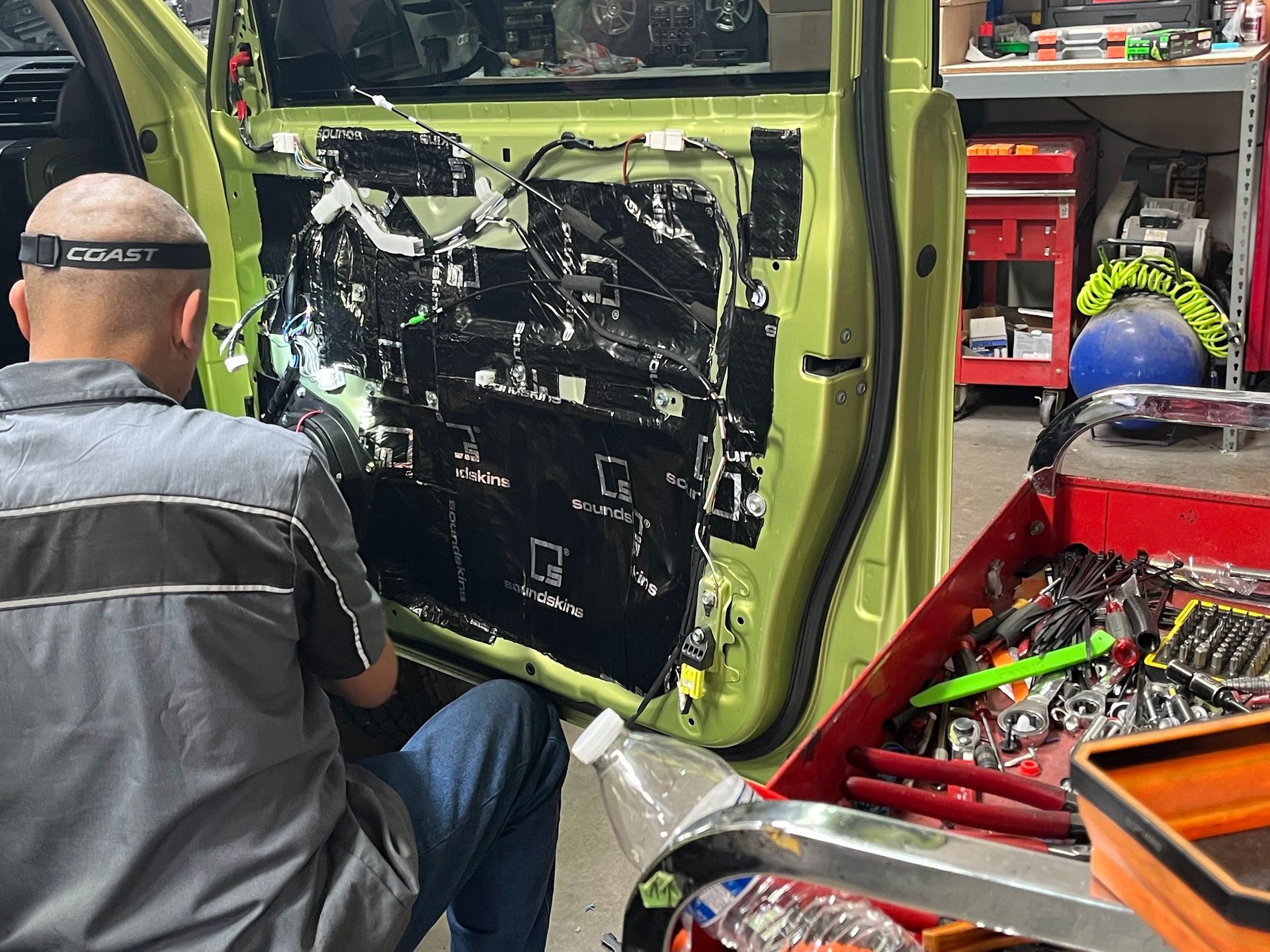 Mechanic working on a car door with soundproofing material applied. Inside a garage, tools in an open toolbox.