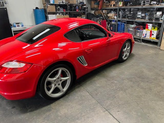Red Porsche Cayman coupe parked inside a garage with shelves of equipment in the background.