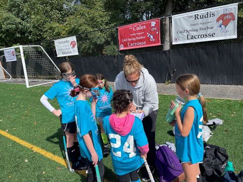 Youth soccer team huddles on the field around a coach near the sideline, with bags and a goal in the background.