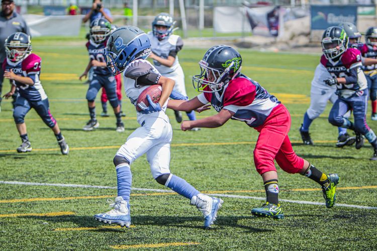 Youth football players in red and blue uniforms tackle a runner in white on a grassy field