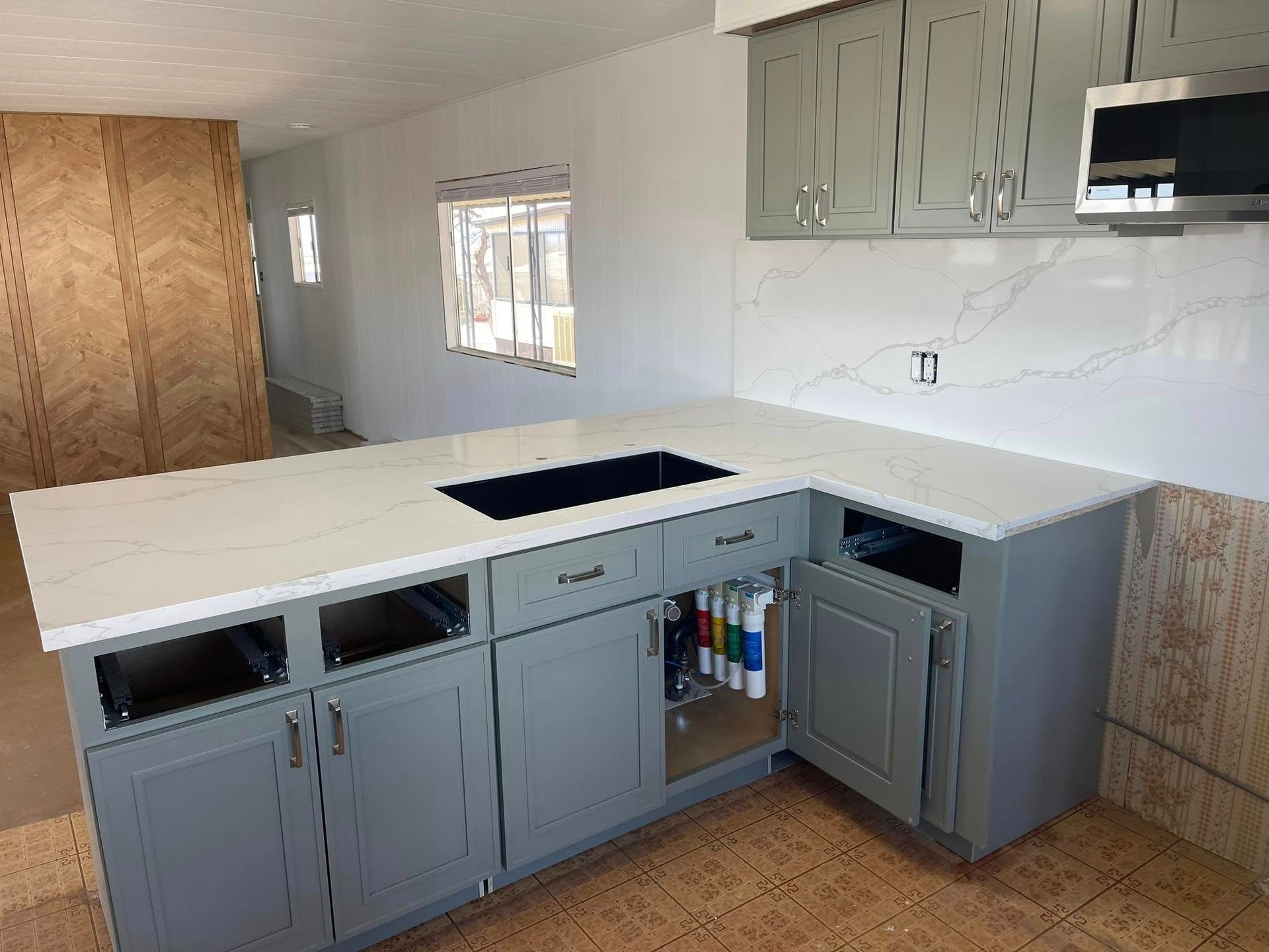 Grey kitchen cabinets with white countertop and backsplash; sink, cooktop, and island.