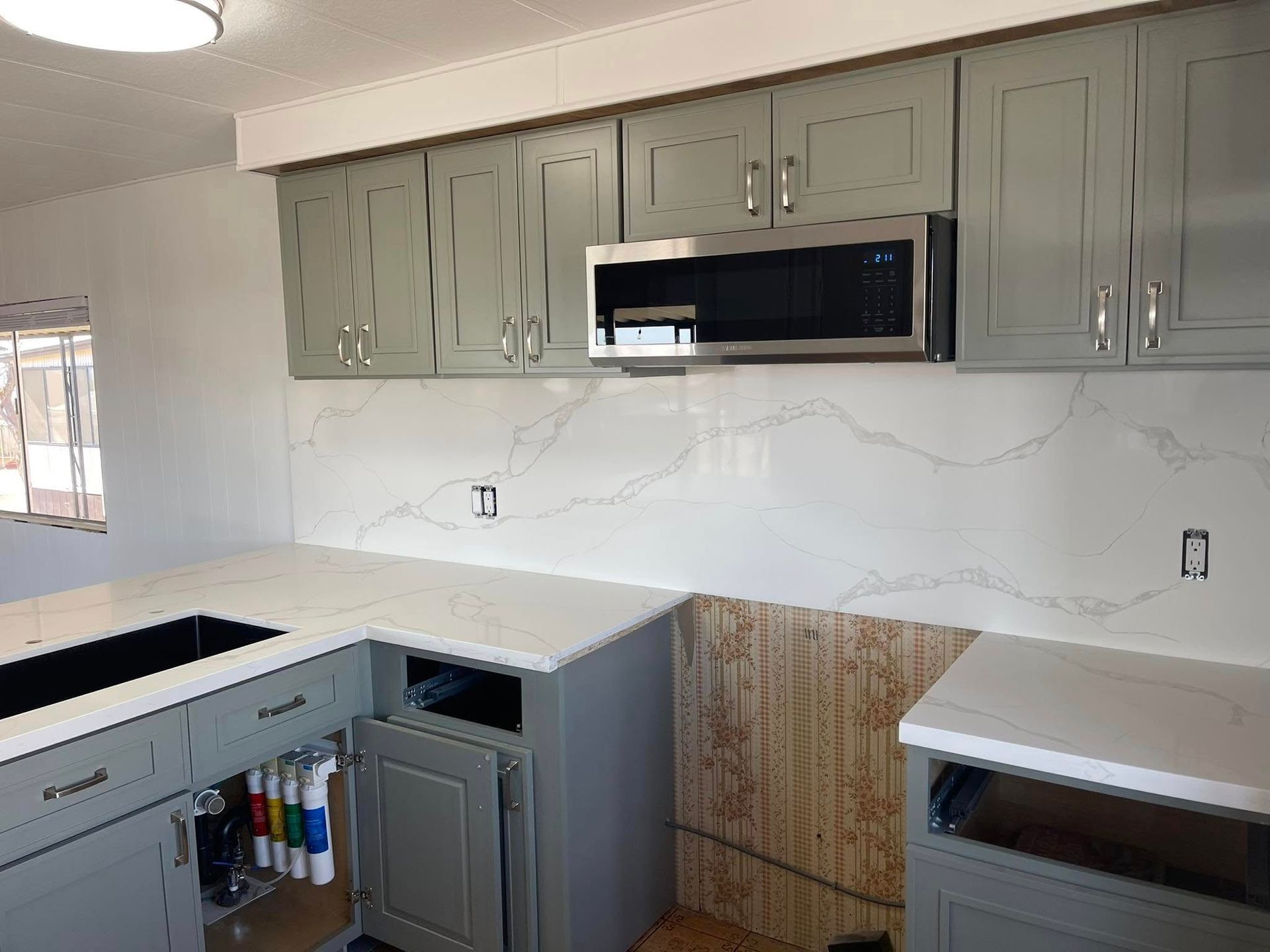 Gray kitchen cabinets and countertops, white backsplash, and stainless steel microwave.