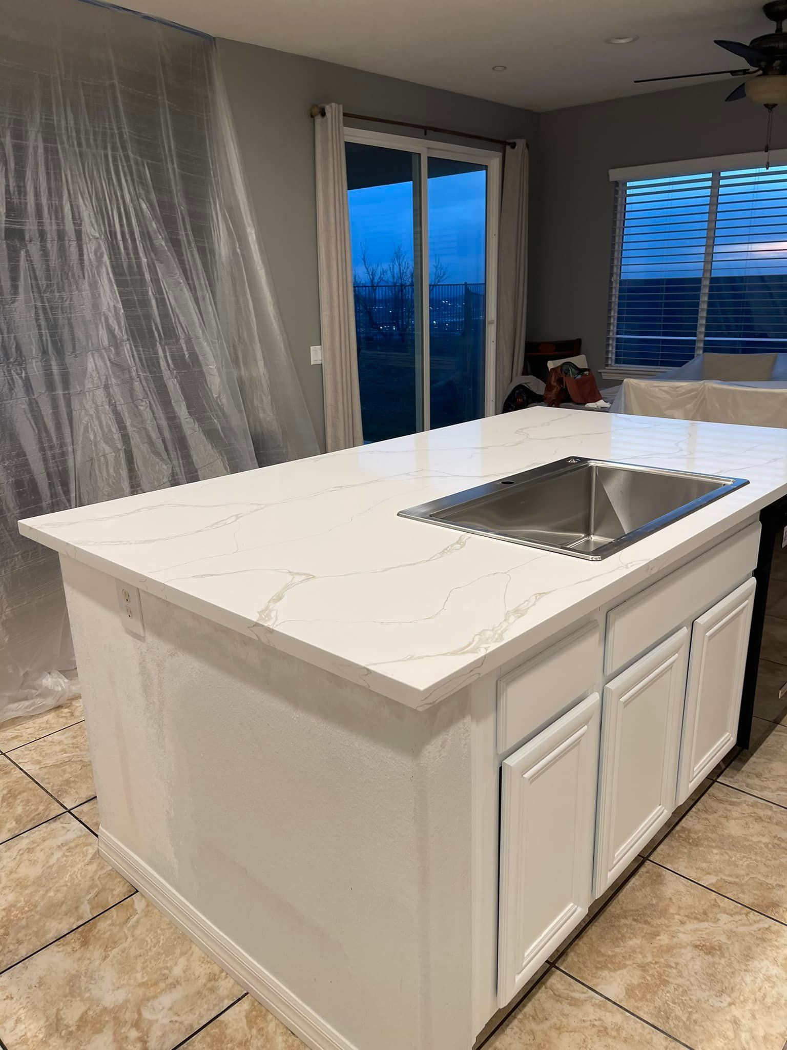 White kitchen island with quartz countertop and sink, white cabinets, tile floor, and sliding glass door.