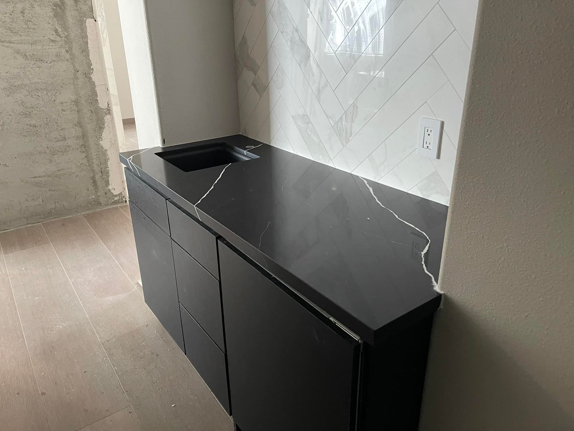 Black kitchen countertop with integrated sink, drawers, and backsplash in an unfinished room.
