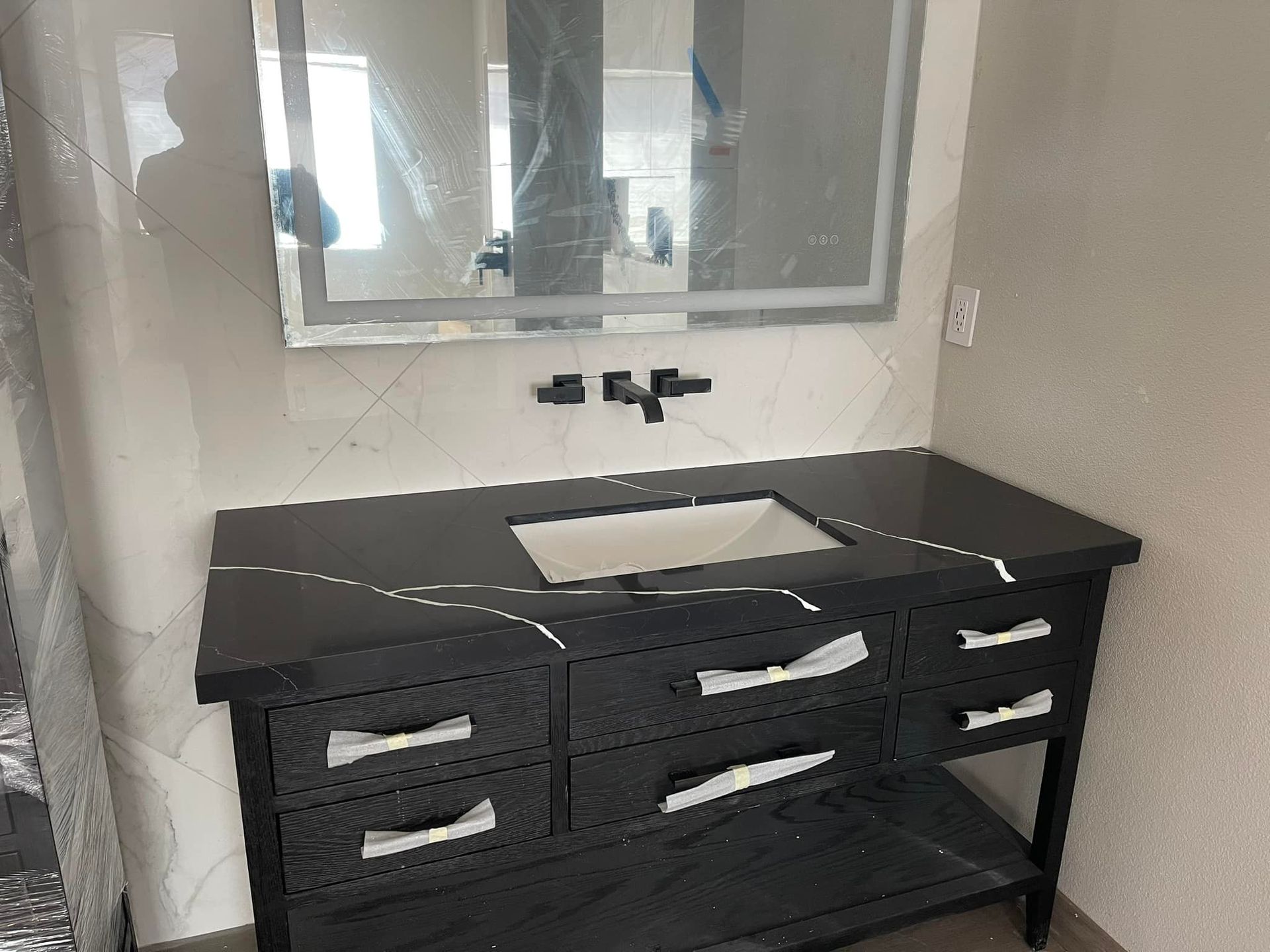 Black vanity with white countertop, sink, and drawers. Wall-mounted black faucet, mirror, and tiled backsplash.