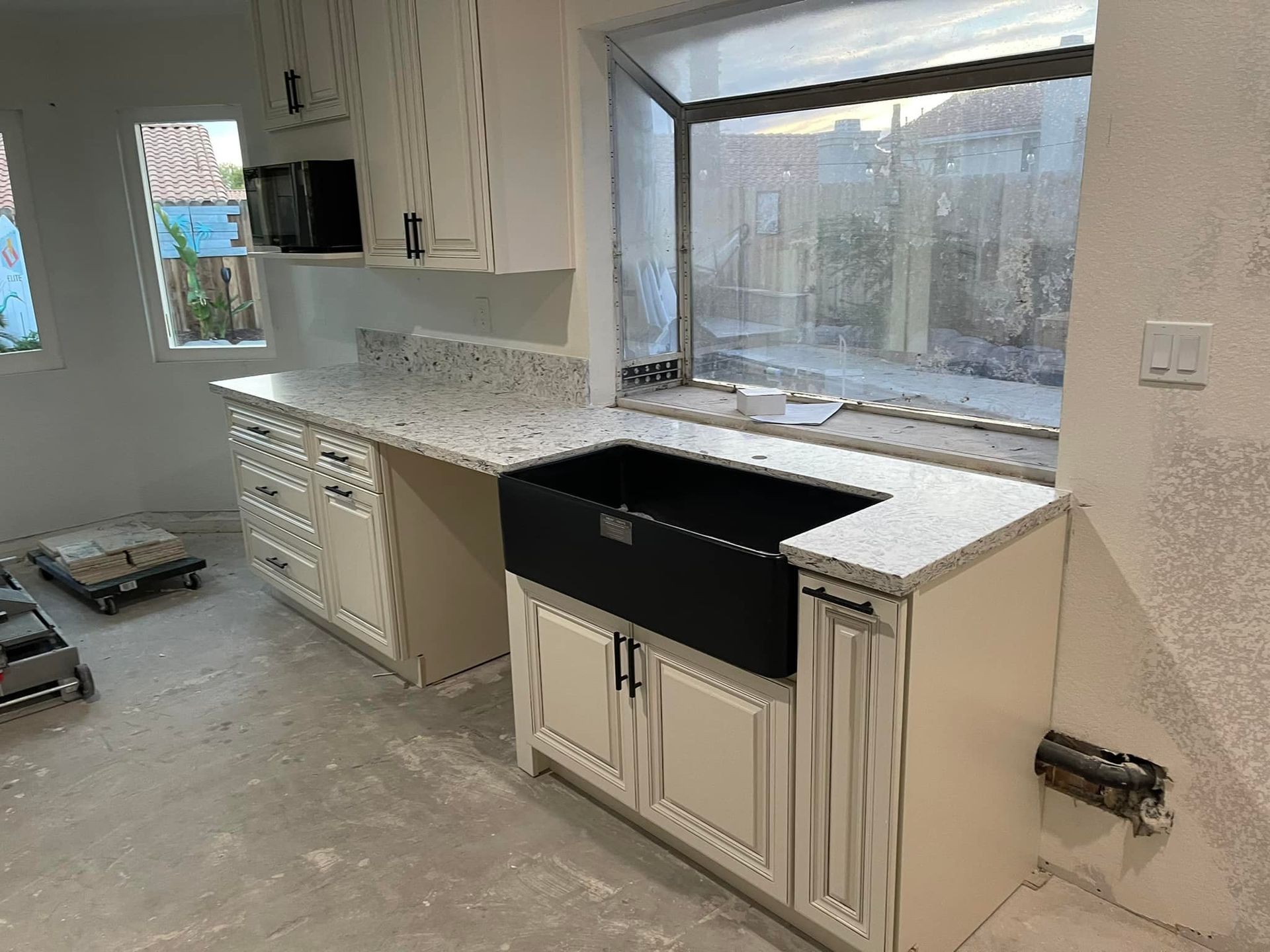 Cream-colored kitchen cabinets with a granite countertop, black farmhouse sink, and a window overlooking a neighborhood.