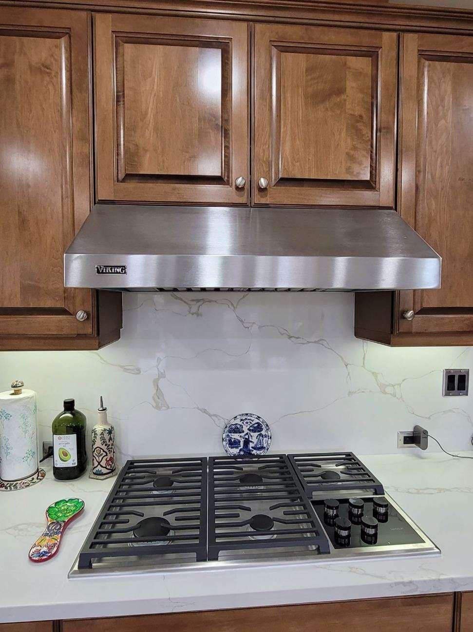 Kitchen with wooden cabinets, stainless steel range hood, marble backsplash, gas stove, and countertop with decor.