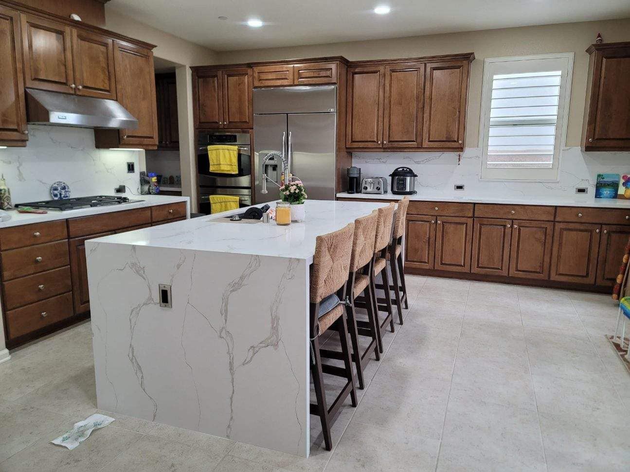Kitchen with brown cabinets, white countertops, island with bar stools, stainless steel appliances.