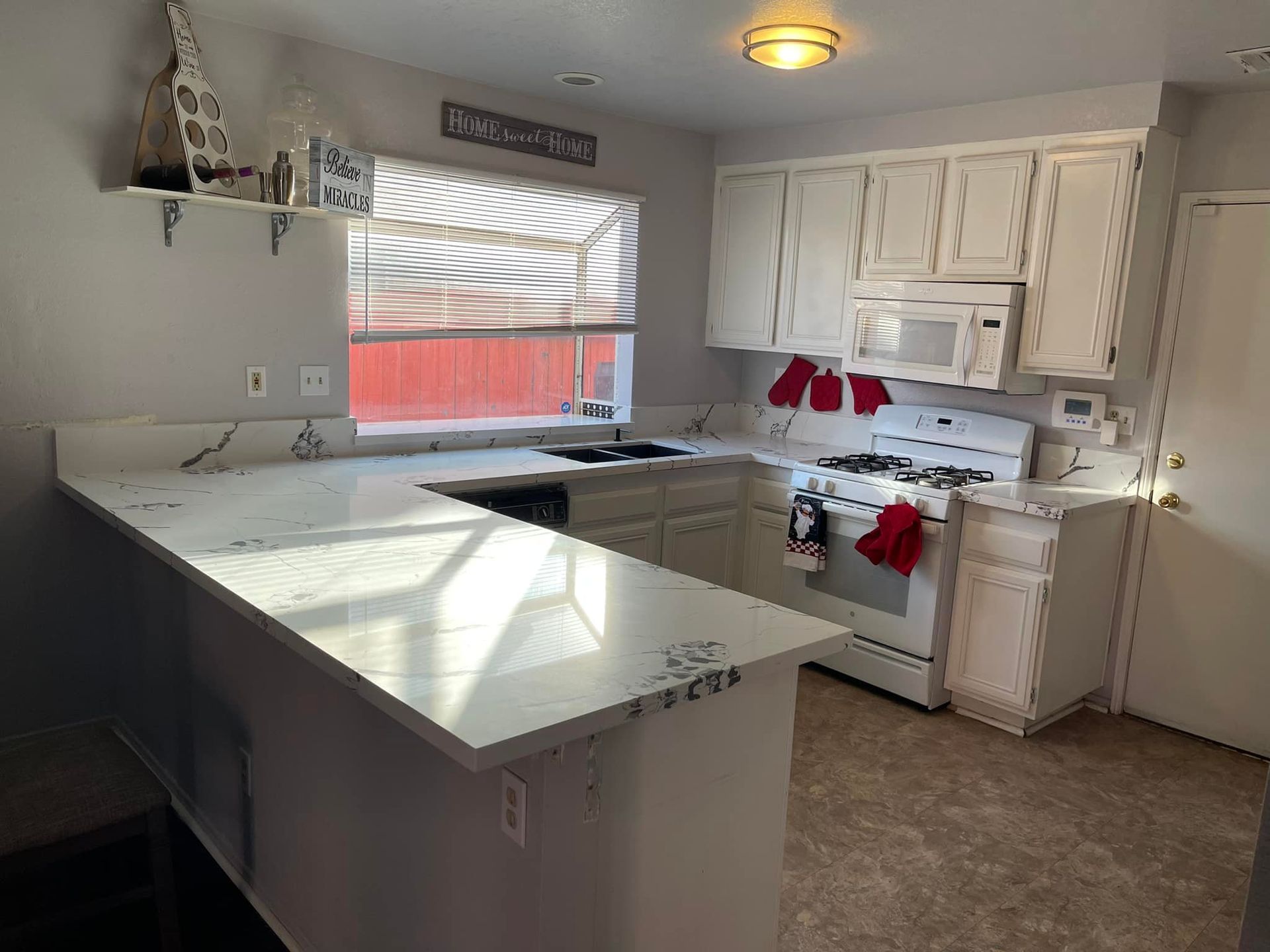 White kitchen with countertop, cabinets, and appliances. Sunlit, window with blinds. Red accents.
