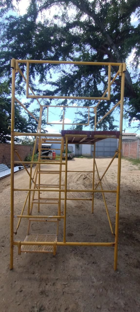 Un andamio amarillo se alza al aire libre, con árboles y un edificio de fondo.