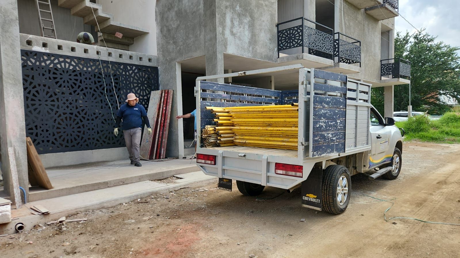 Camión cargado con andamios amarillos, estacionado frente a un edificio en construcción. Un hombre camina hacia la entrada.