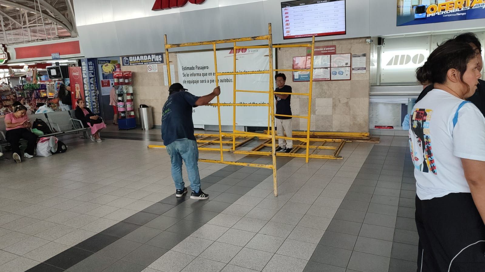 Hombre montando un andamio amarillo alrededor de una pizarra blanca en una zona pública con gente y tiendas.