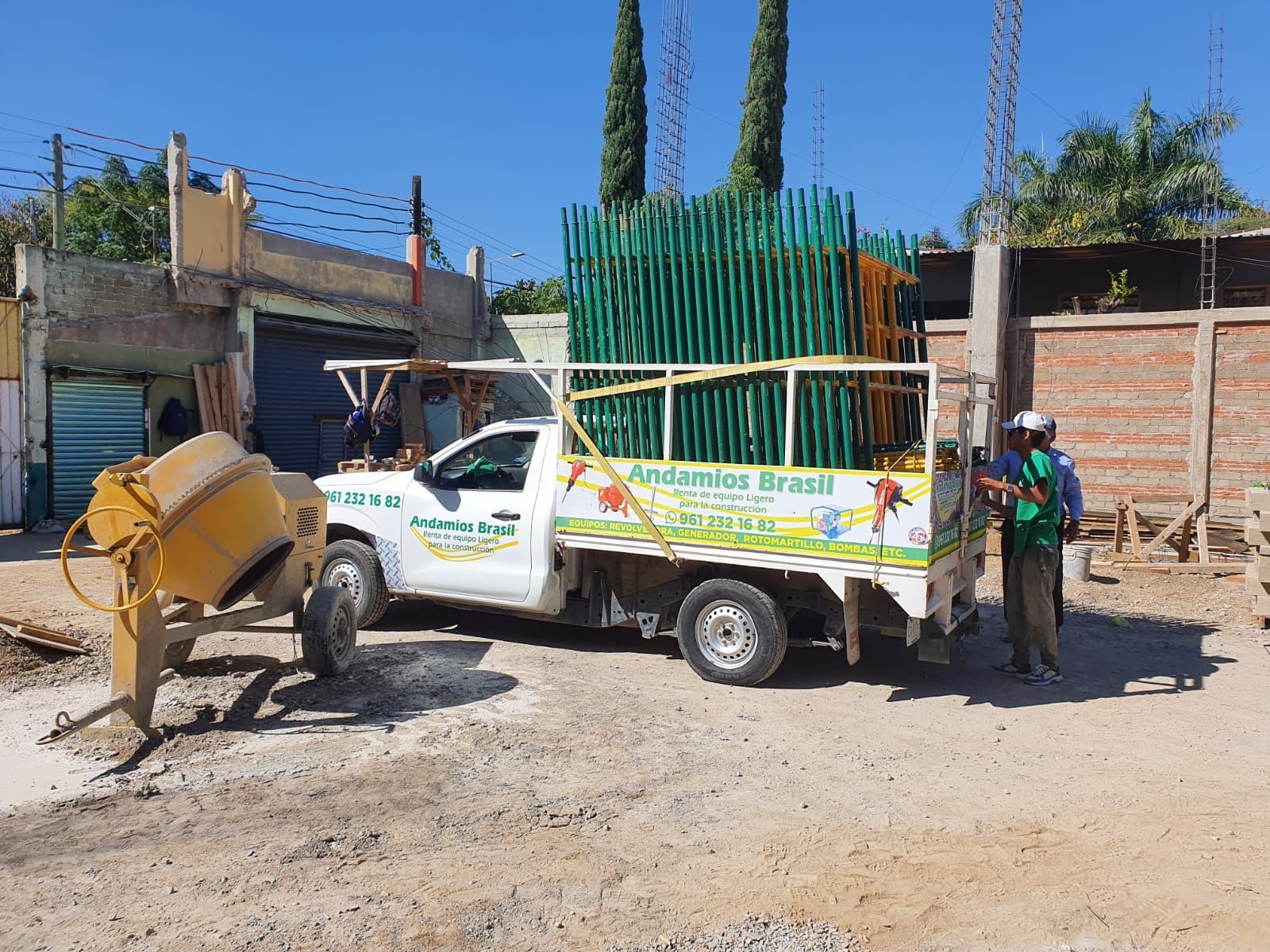 Camión blanco cargado con andamios verdes y una hormigonera, estacionado en un camino de tierra junto a una persona.