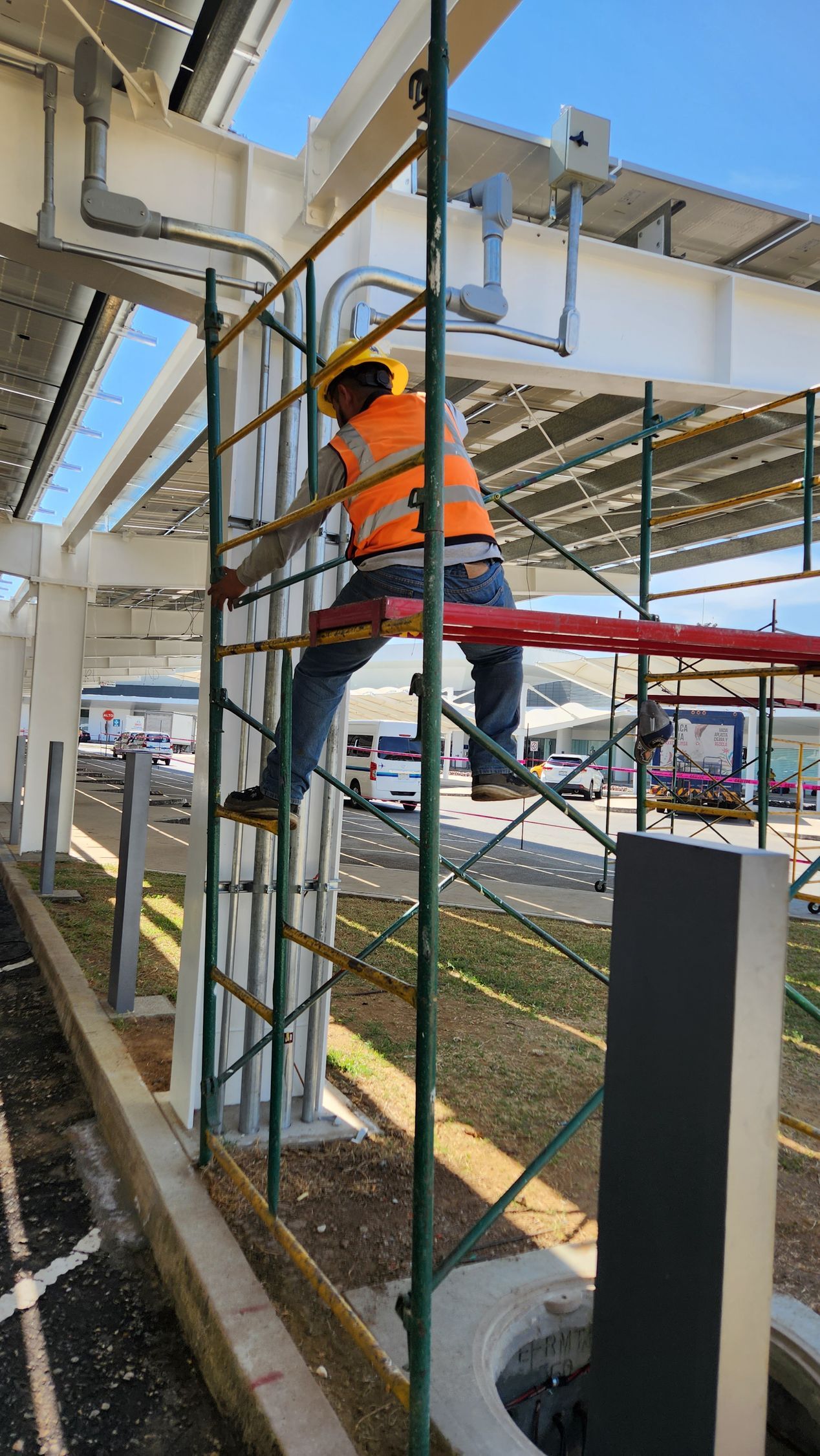 Obrero de la construcción sobre un andamio, instalando tuberías bajo una estructura de paneles solares.