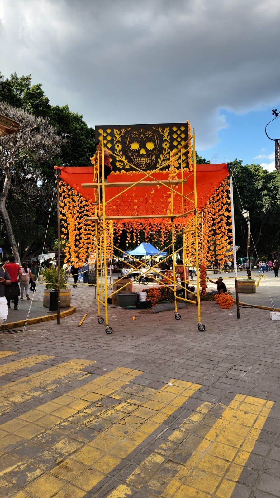 Estructura de andamio naranja decorada con motivos del Día de Muertos, con diseño de calavera, en una plaza pública.
