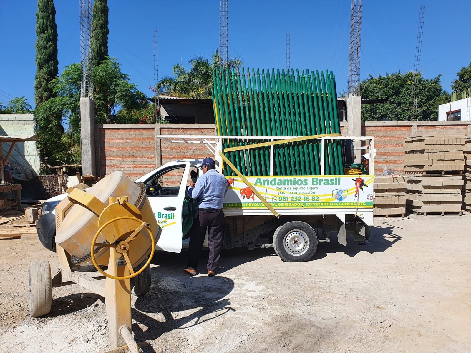Hombre subiendo a un camión cargado con andamios verdes y una hormigonera en una obra en construcción.