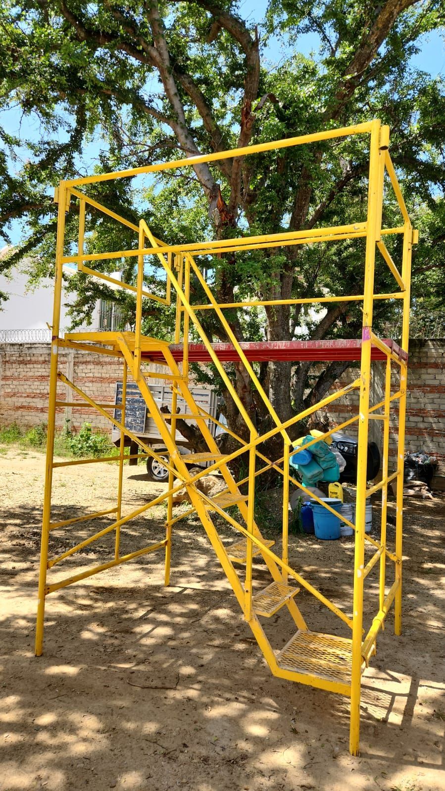 Andamio metálico amarillo con escaleras, instalado al aire libre, apoyado contra un árbol.