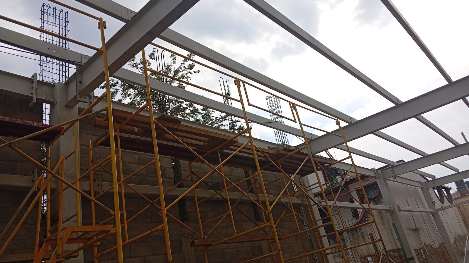 Obra en construcción con andamios amarillos, muros de hormigón y vigas de acero contra un cielo nublado.