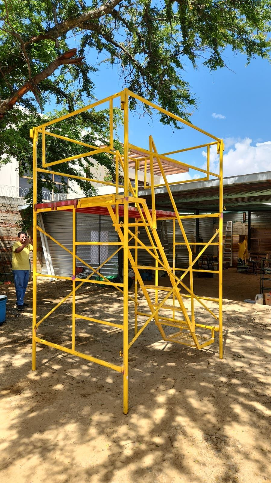 Estructura de andamio amarilla con escaleras, persona de pie cerca, al aire libre.