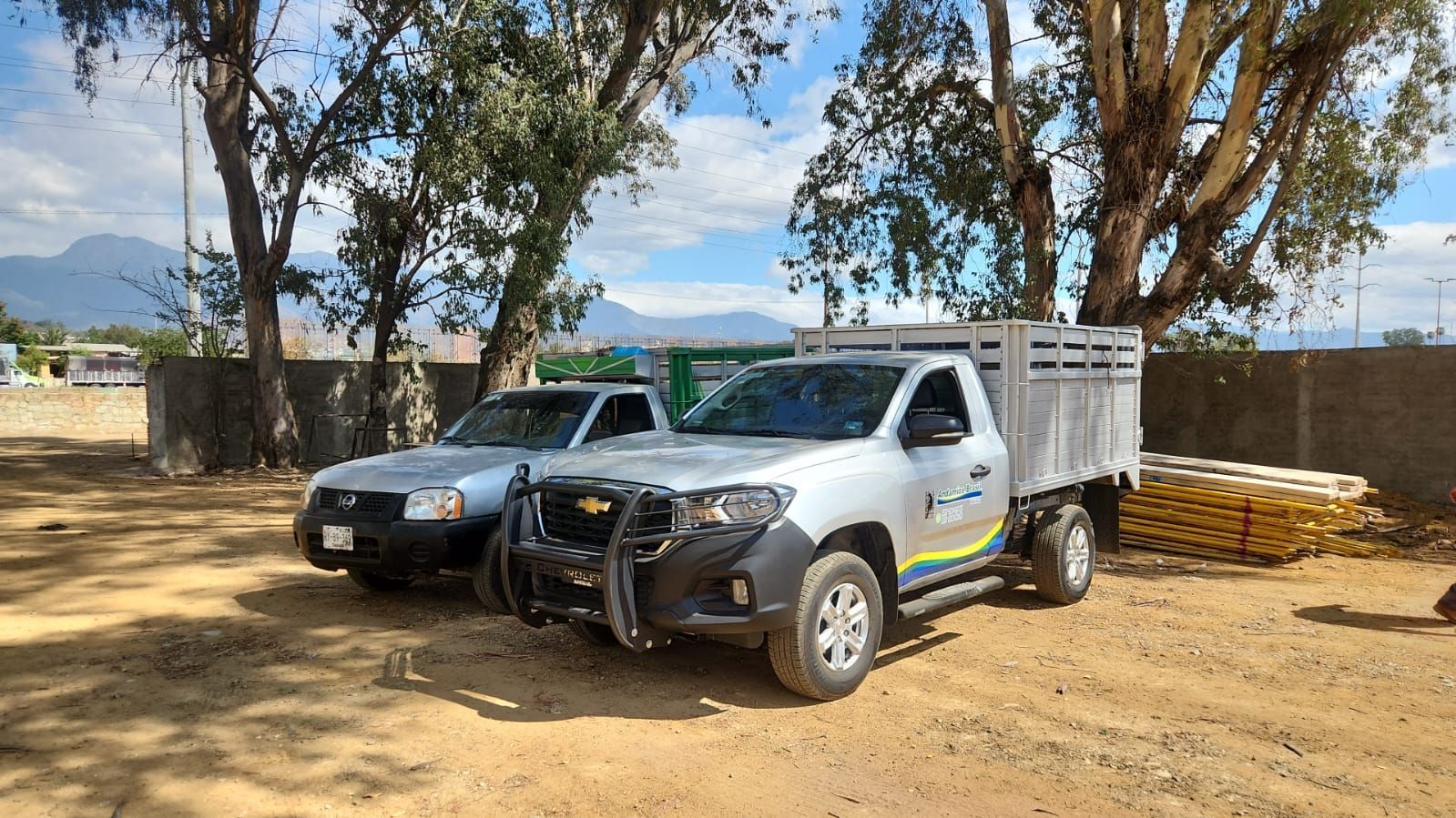 Dos camionetas estacionadas al aire libre sobre tierra. Una camioneta Chevy gris con caja de carga y un auto plateado se encuentran frente a una pared y árboles.