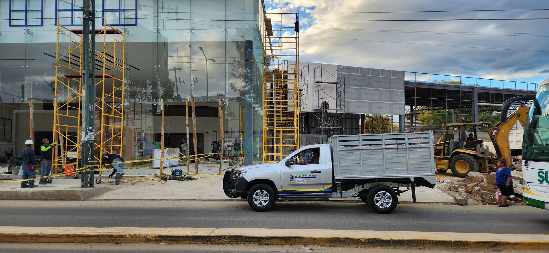Obra en construcción con una camioneta plateada circulando por la calle. Se ven andamios, obreros y maquinaria.