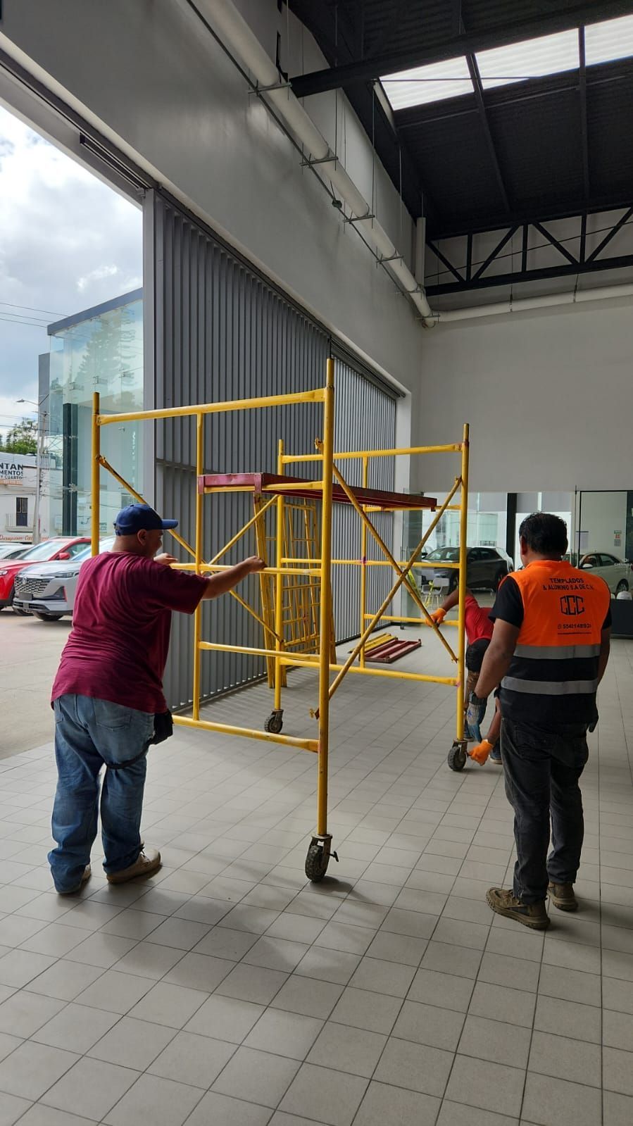 Dos trabajadores maniobran un andamio amarillo dentro de un edificio con una gran puerta abierta.