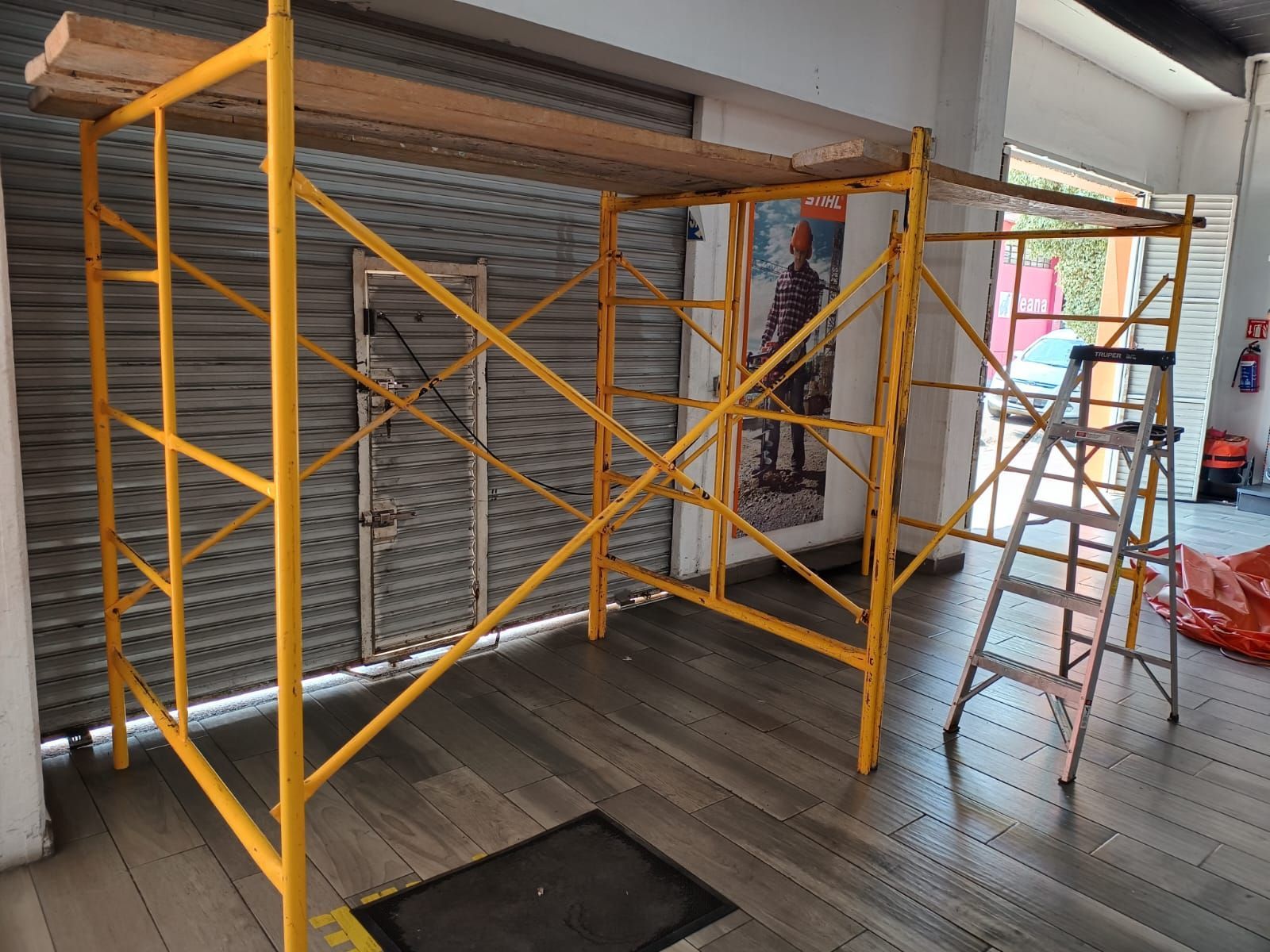 Andamio amarillo con tablones de madera, dentro de un edificio. Una escalera está al lado.