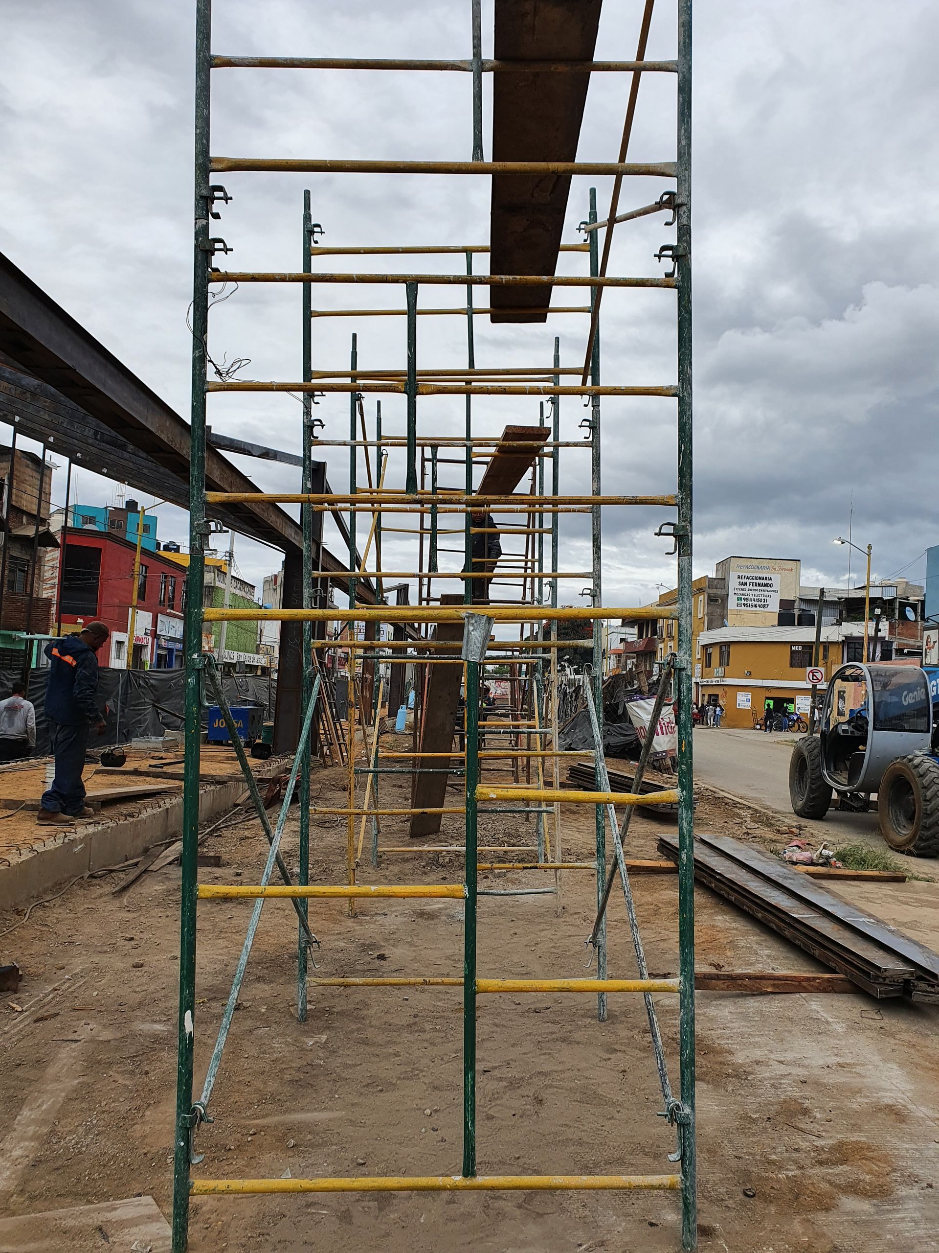 Andamios instalados en una obra en construcción. Al fondo se ven obreros y edificios. Cielo nublado.