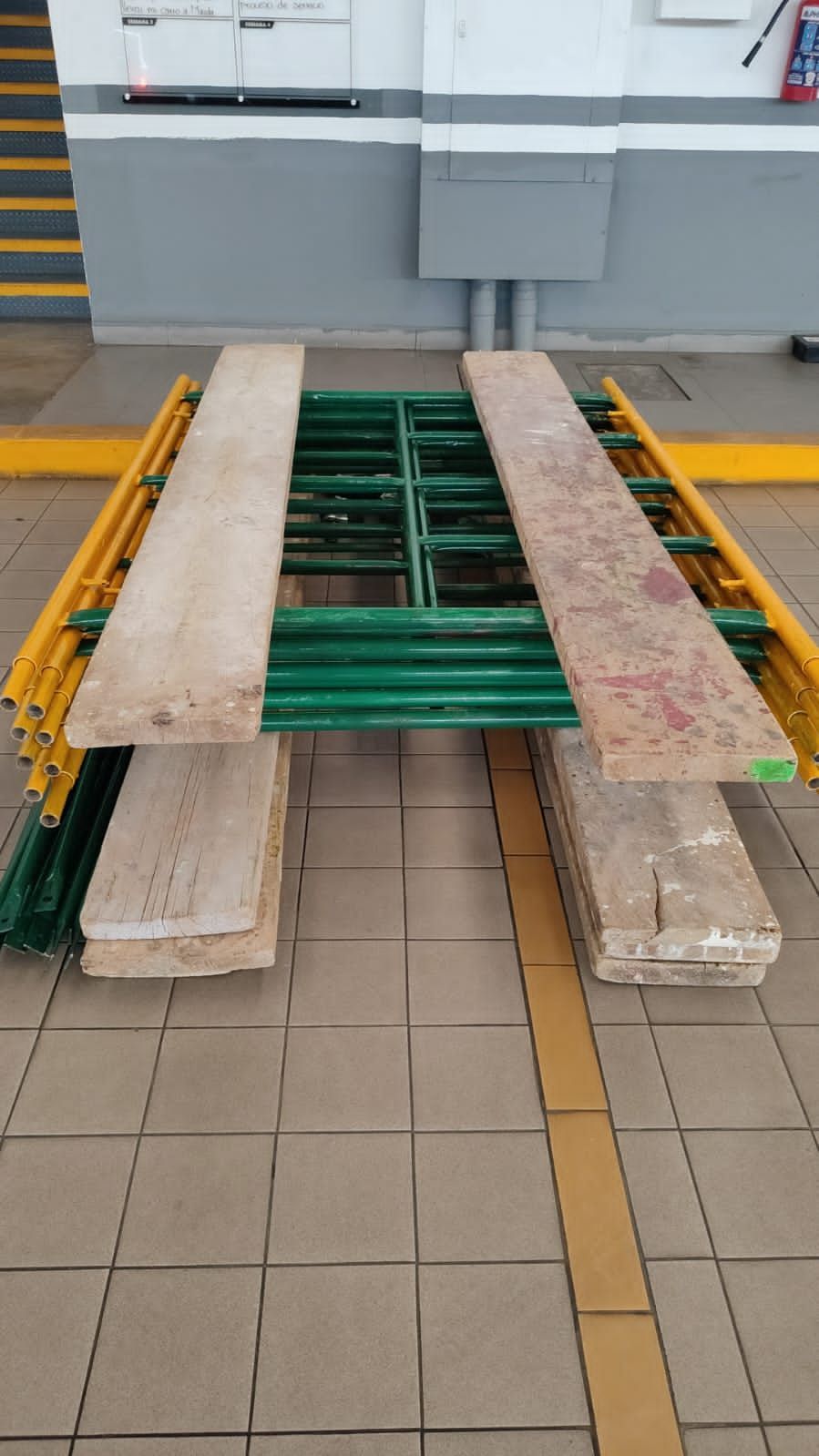 Andamio con tablones de madera sobre suelo de baldosas en el interior de un edificio. Estructura metálica amarilla y verde.