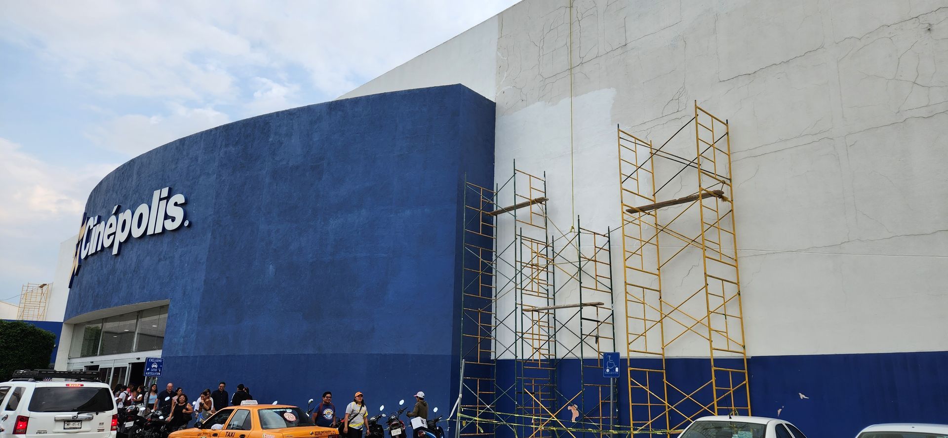 Vista exterior de un cine Cinépolis, fachada azul y blanca, con andamios, gente y vehículos delante.