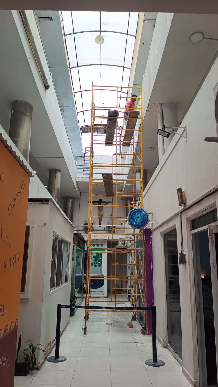 Una persona subiéndose a un andamio repara una claraboya en un pasillo interior lleno de tiendas.