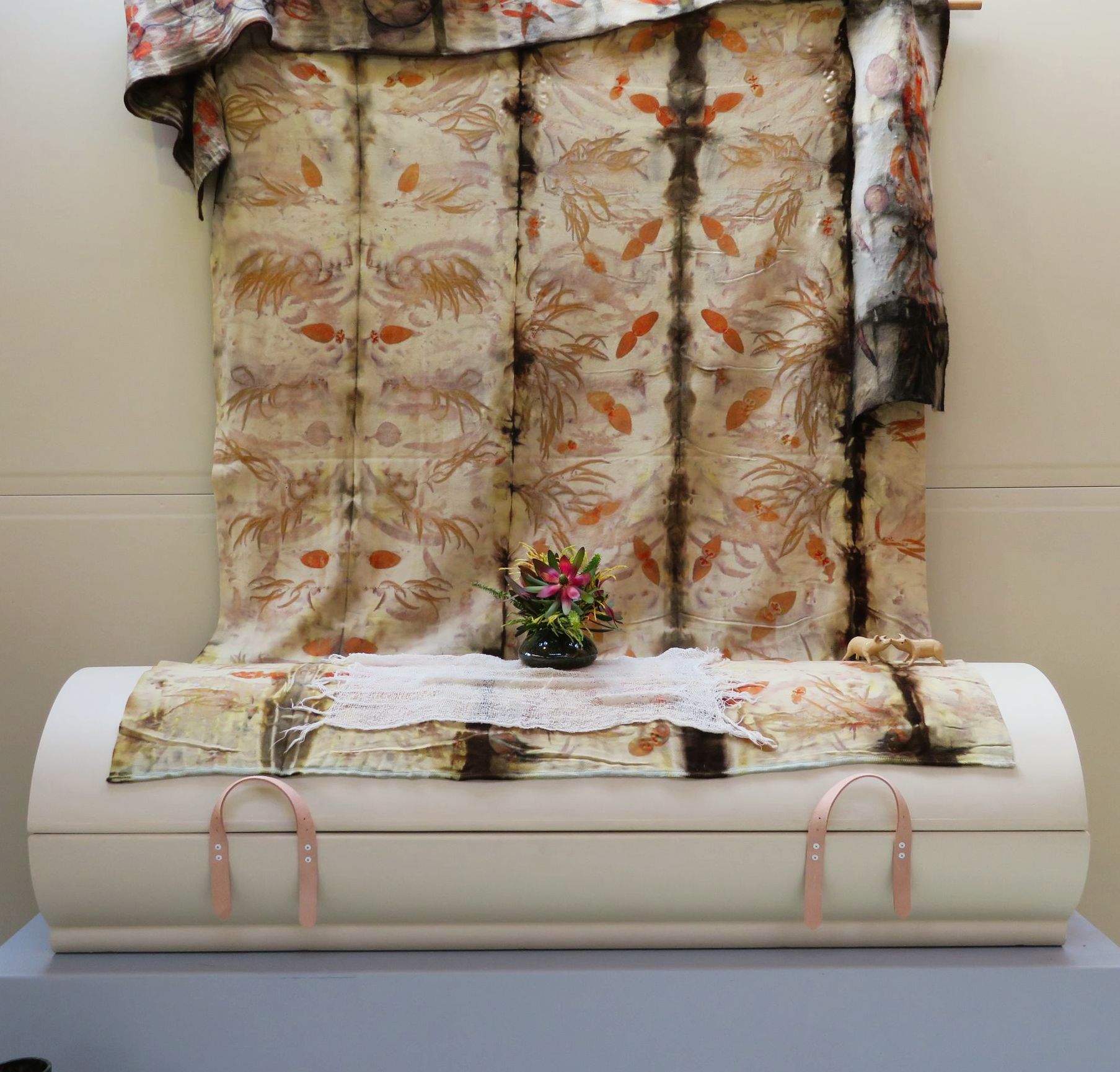 A white coffin covered with a floral pattern wrapped is on a stand in front of a natural printed wall hanging