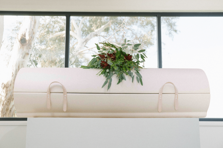 A white coffin with flowers on top of it is sitting in front of a window.