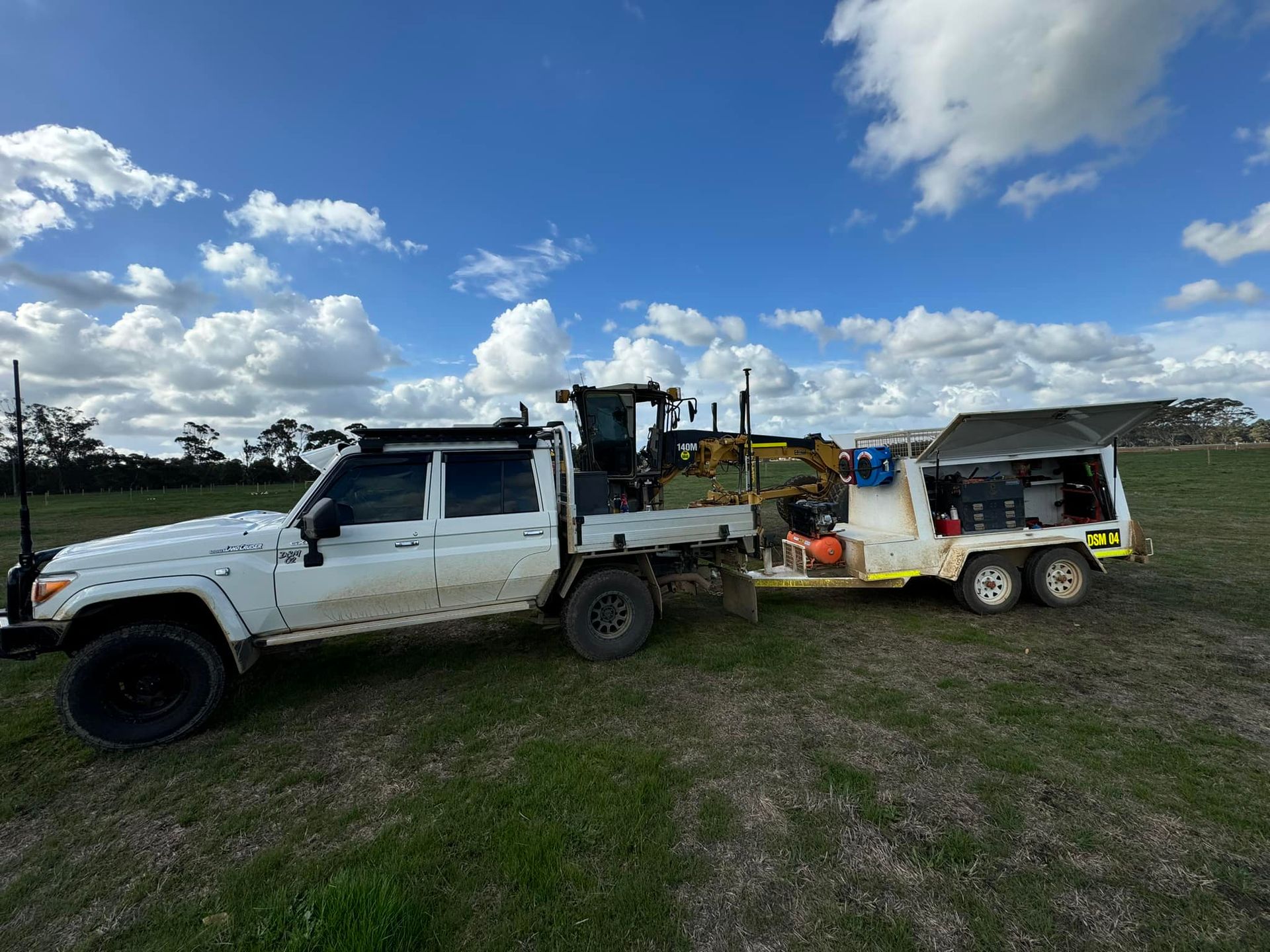 White truck with a mini excavator on its flatbed and a trailer on a grassy field under a cloudy sky.