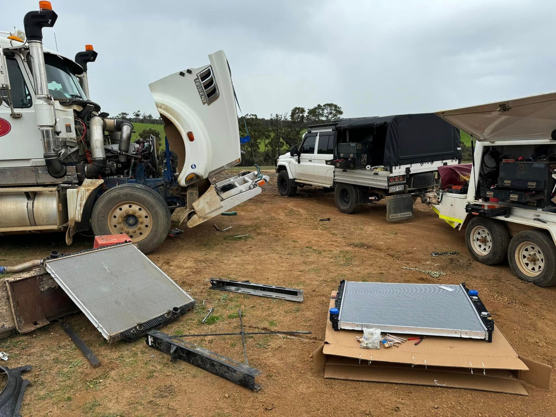 Semi-truck with open hood, radiator on ground, work trucks in rural setting.