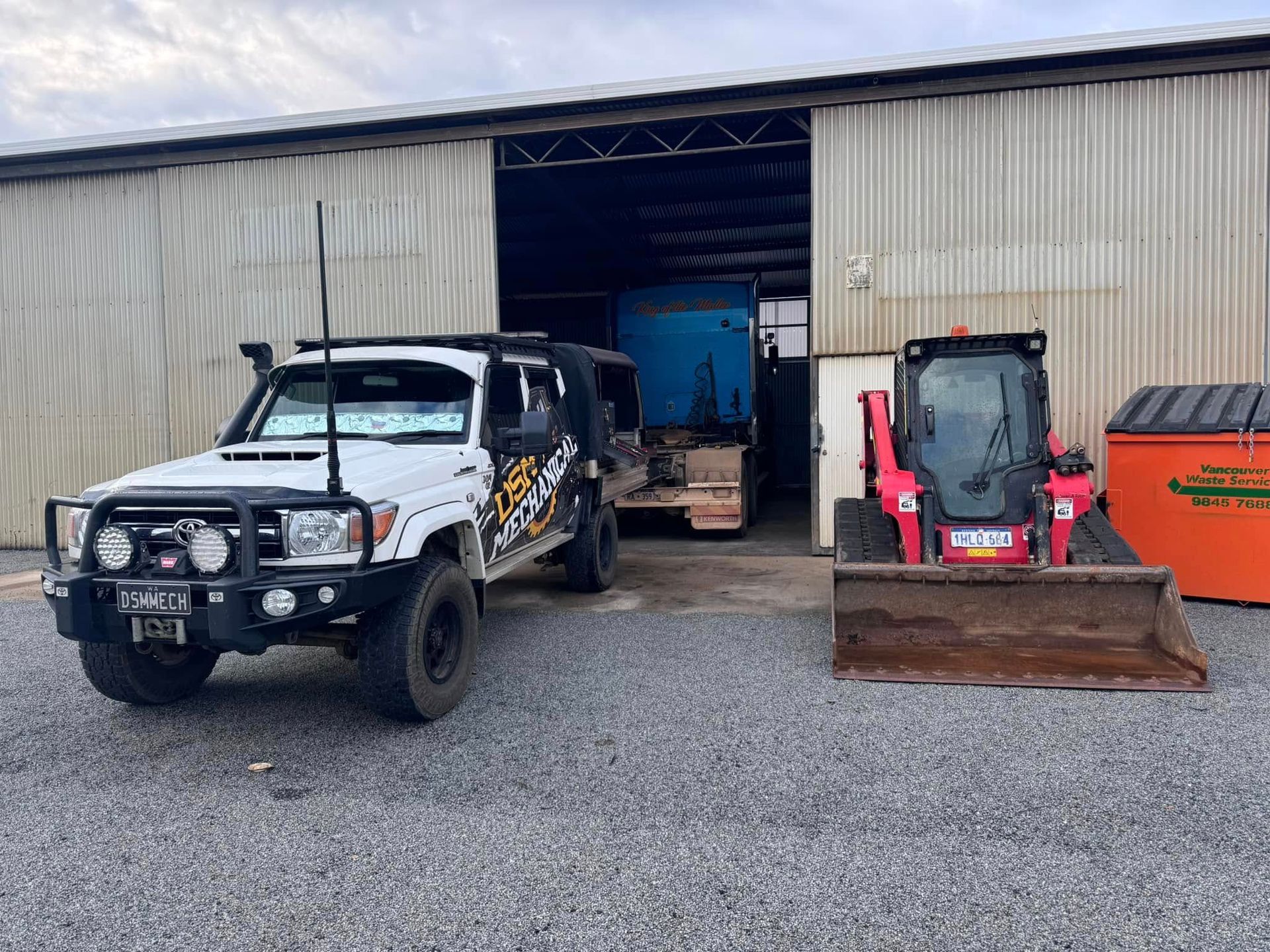 White off-road vehicle and pink skid steer loader parked in front of a large open warehouse.