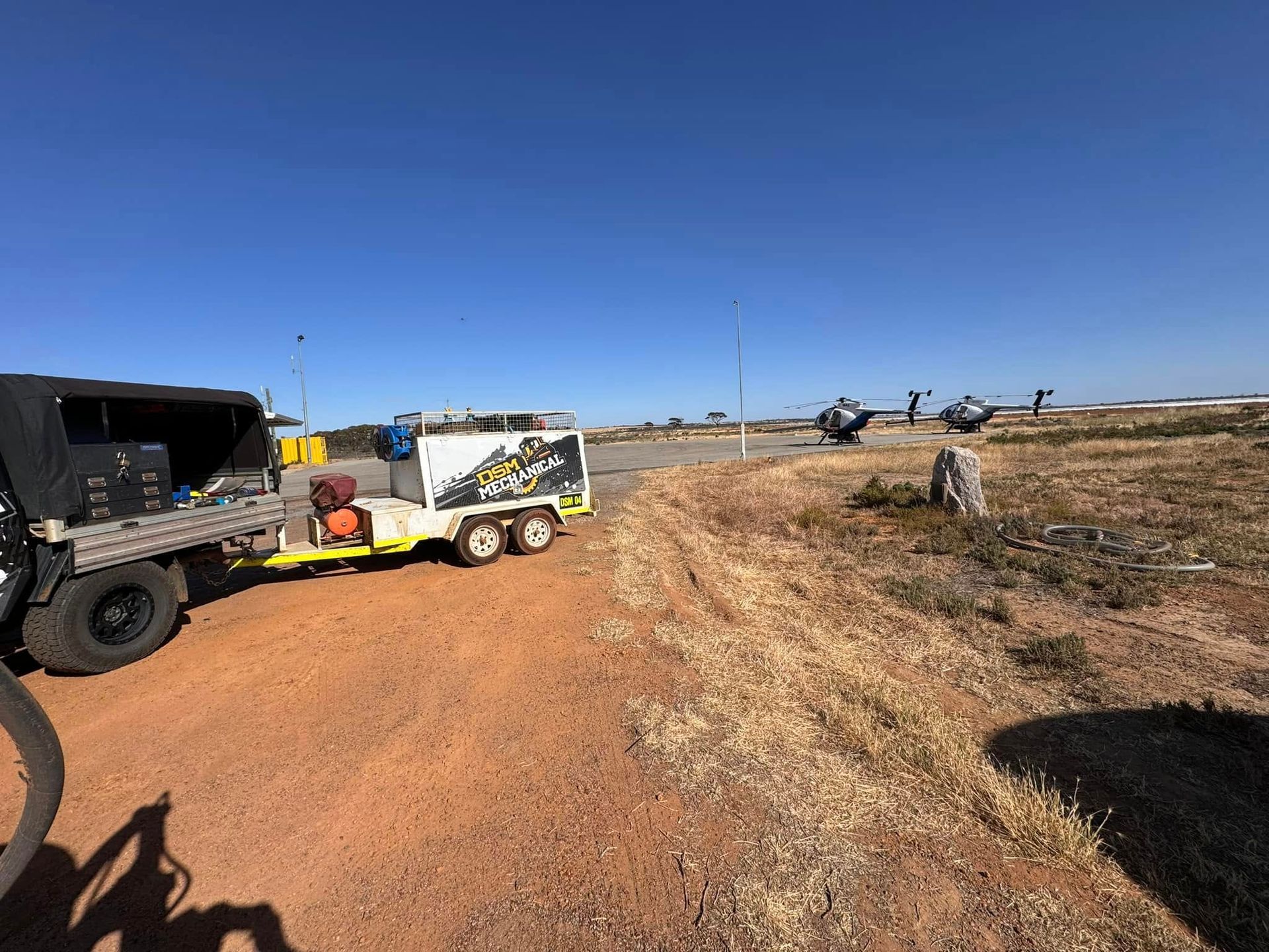 A pickup truck and trailer parked on dirt road near an airfield with helicopters and blue sky.