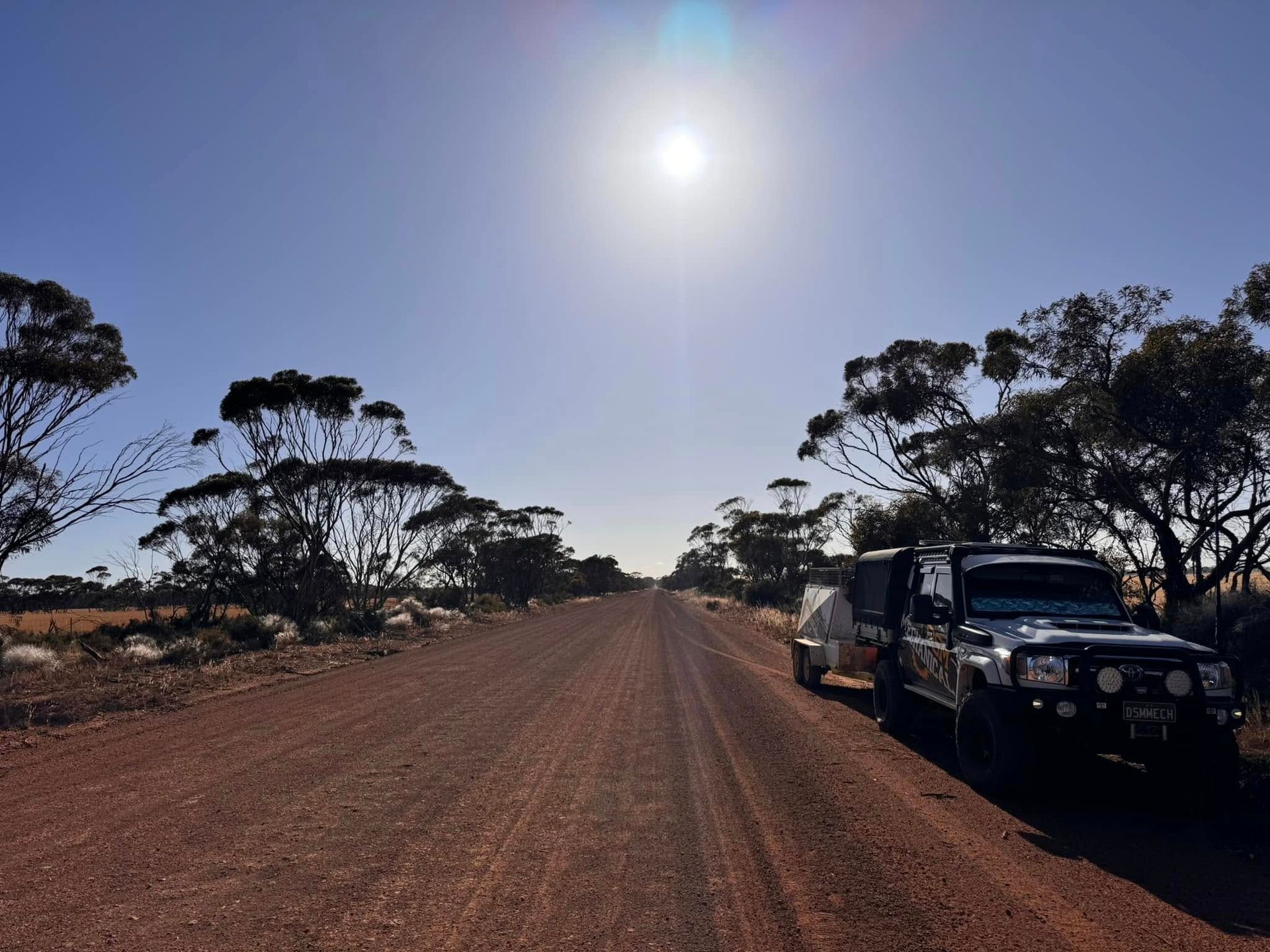 Jeep with trailer parked on a red dirt road under a bright sun, flanked by trees and scrubland.