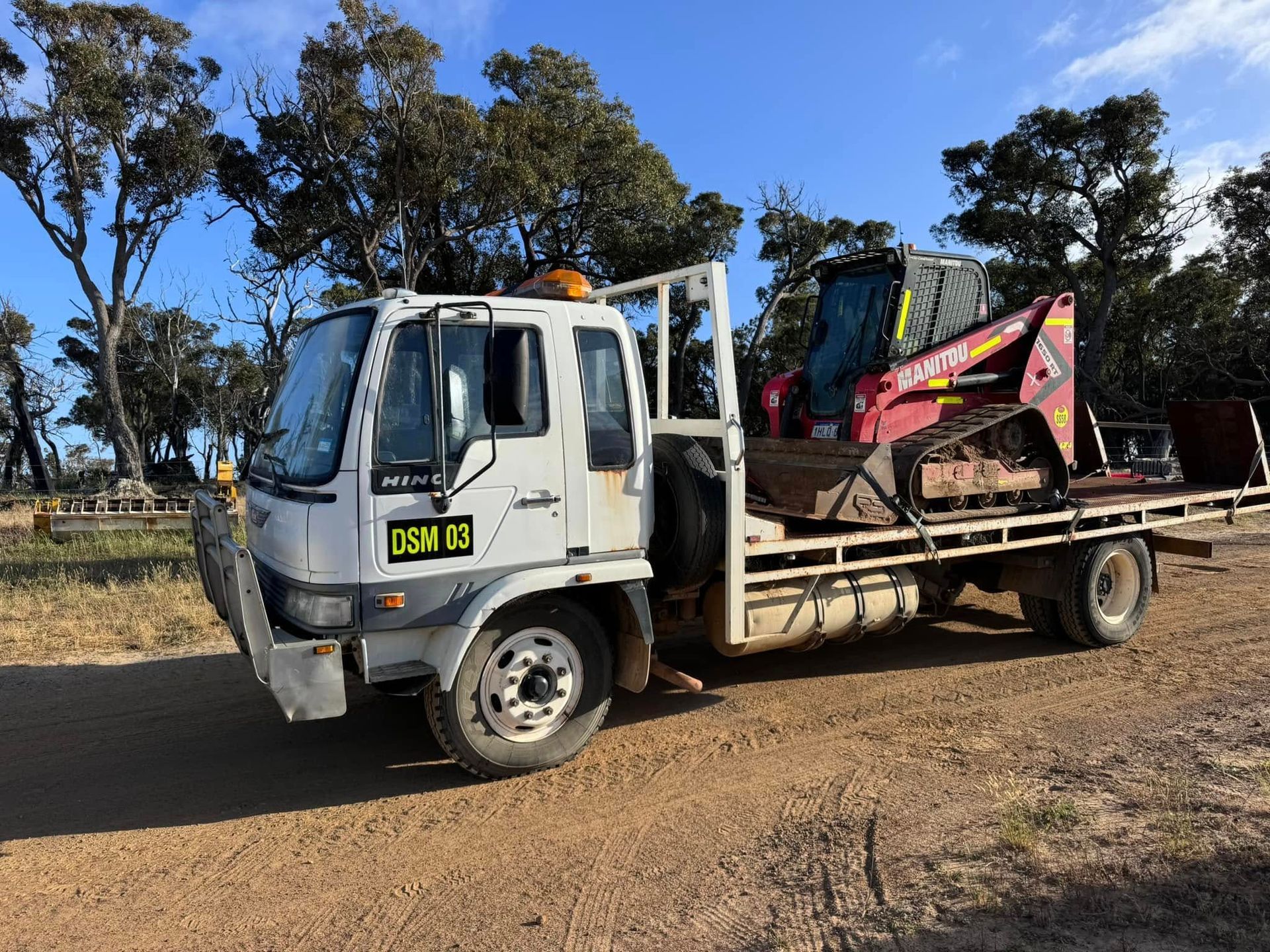 White flatbed truck carrying a red skid steer loader on a dirt road, trees in the background.