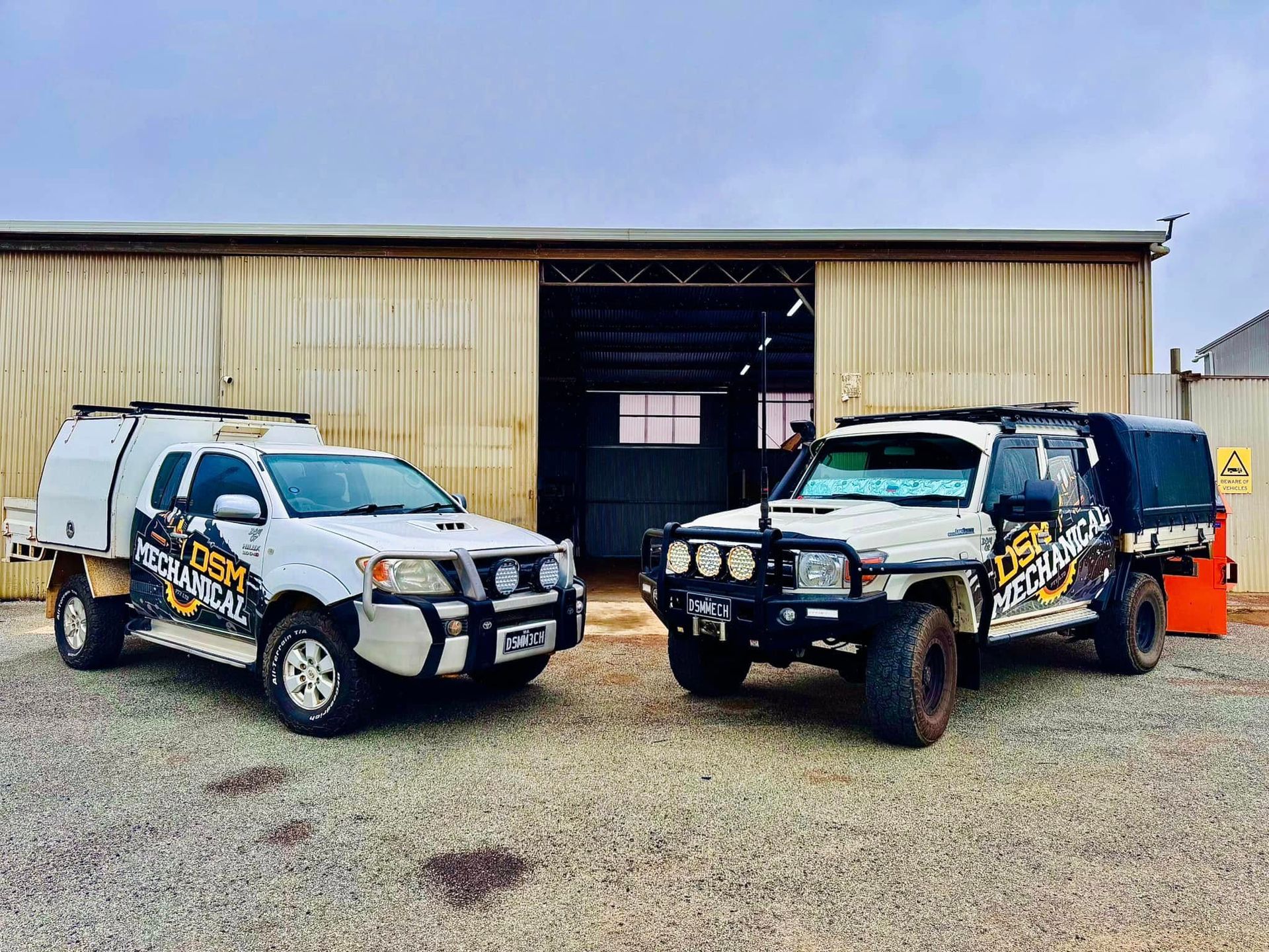 Two white utility trucks with company logos parked in front of a metal building.