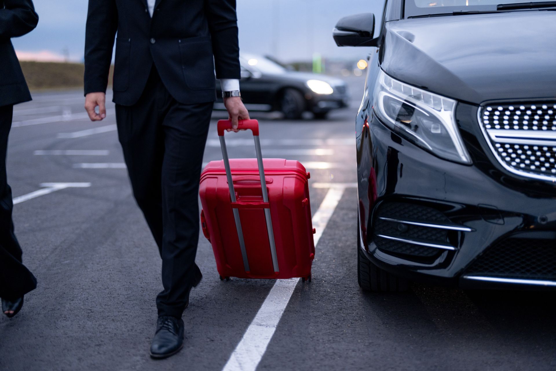 Person rolling a red suitcase beside a black car in a parking lot