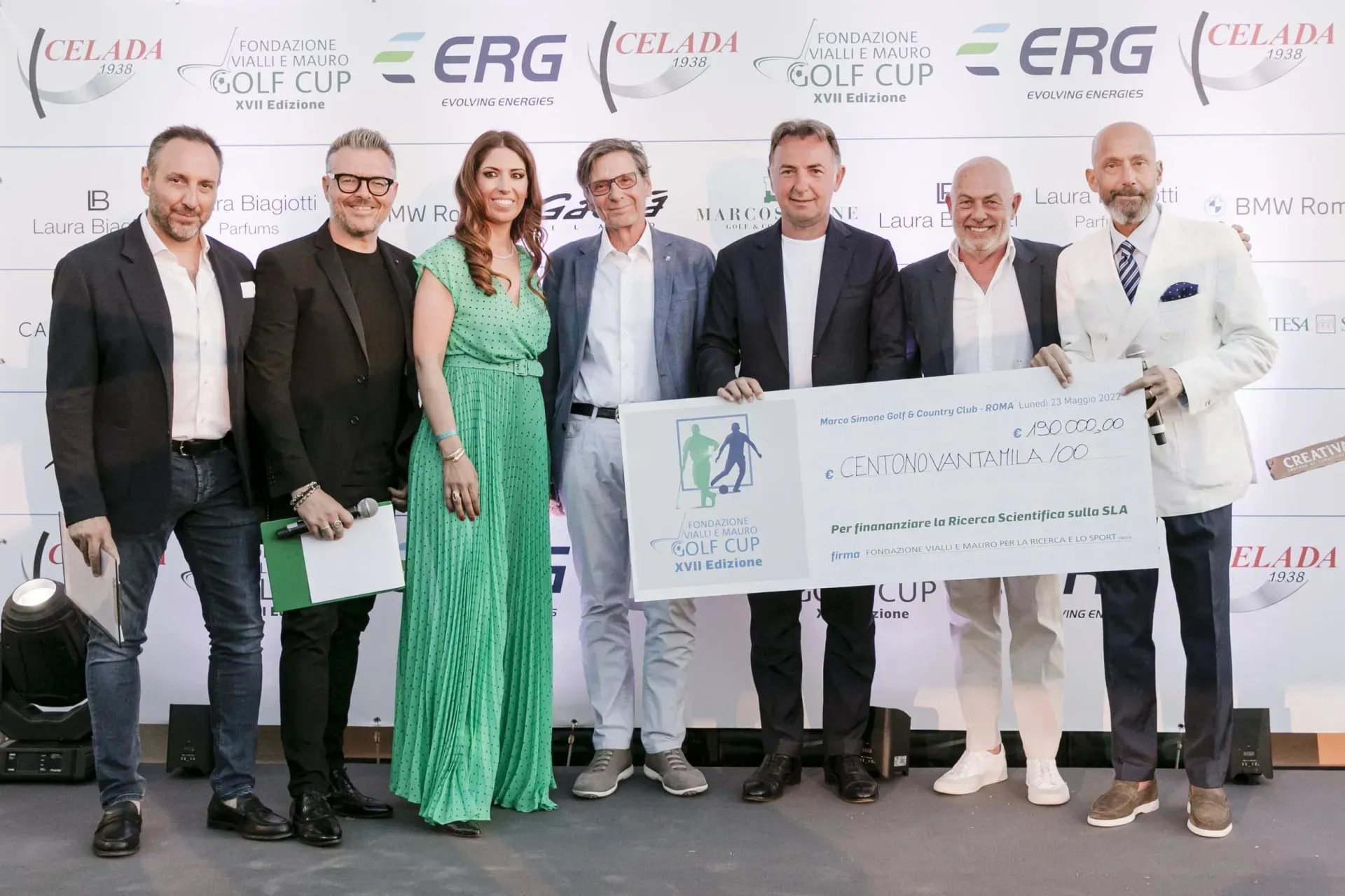 Un gruppo di sette persone tiene in mano un assegno di grandi dimensioni durante un evento all'aperto, probabilmente un torneo di golf.