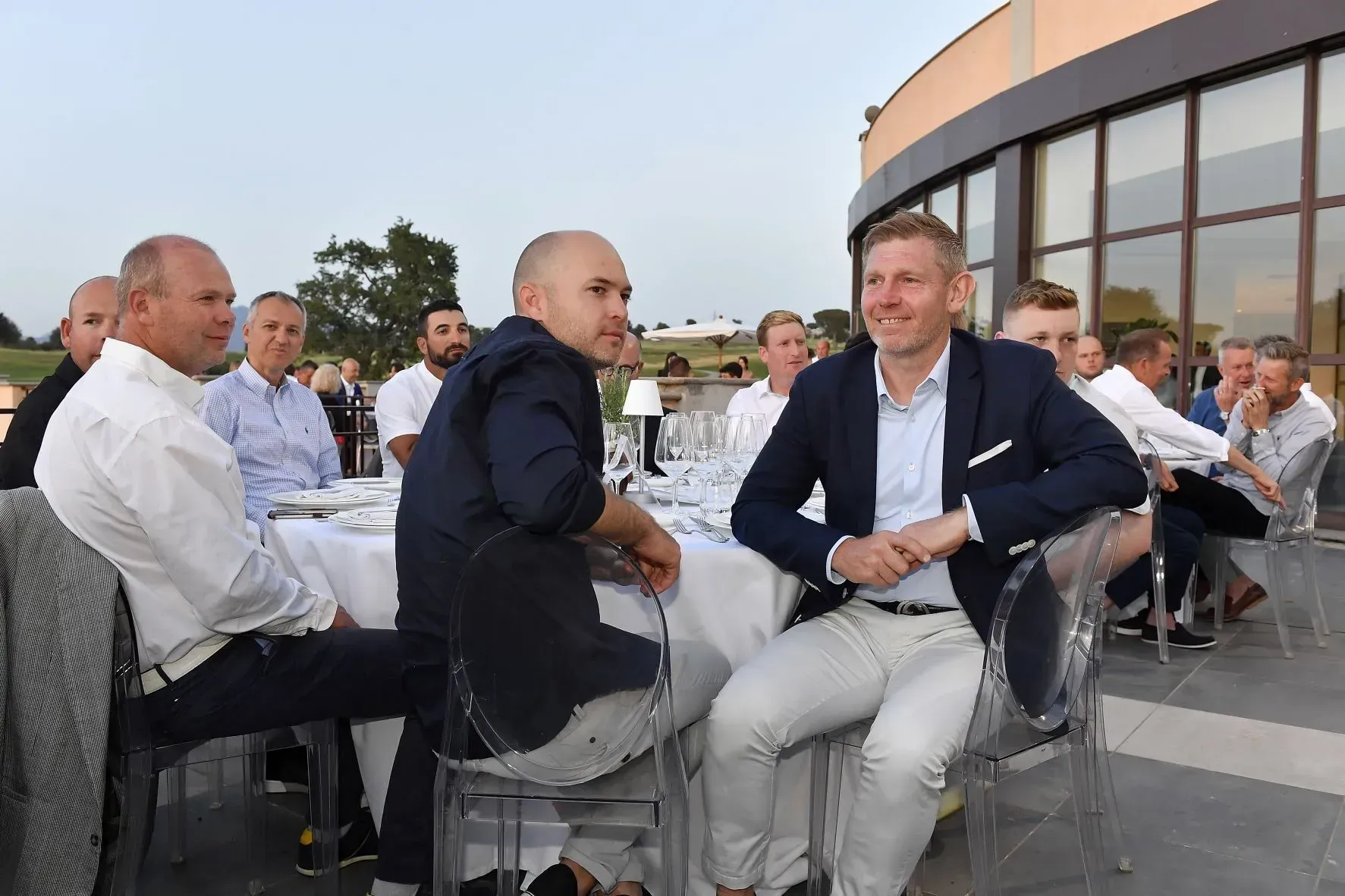Persone sedute a un tavolo all'aperto, durante un evento. Alcune parlano e sorridono.