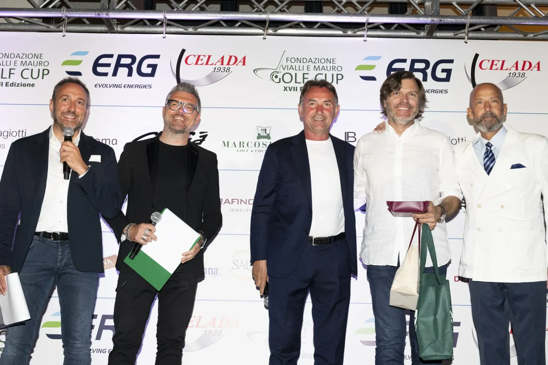 Cinque uomini su un palco con degli oggetti in mano, in posa. Dietro di loro c'è uno sfondo con dei loghi.