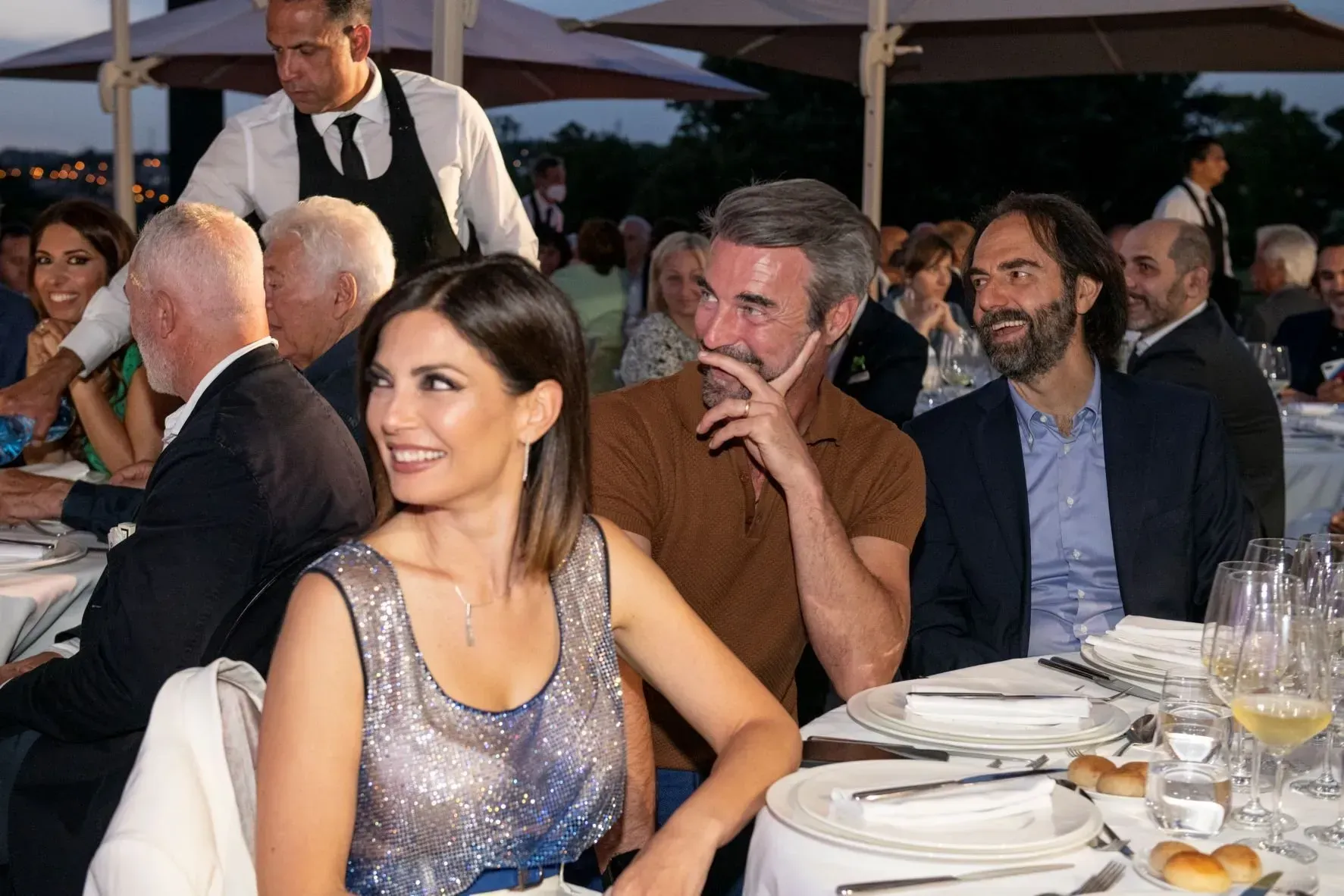 Persone sedute a un tavolo da pranzo all'aperto, sorridenti. Un cameriere serve sullo sfondo.