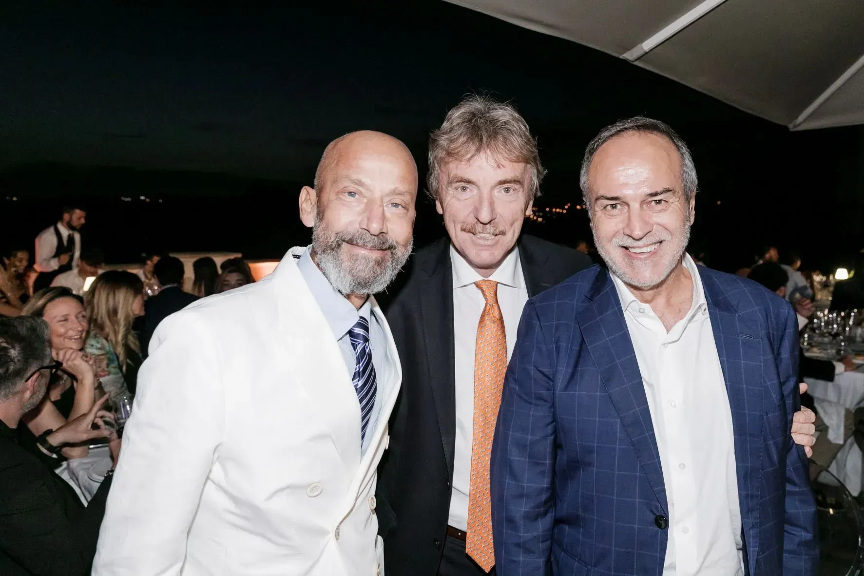 Tre uomini in abiti formali posano all'aperto durante un evento, sorridenti.