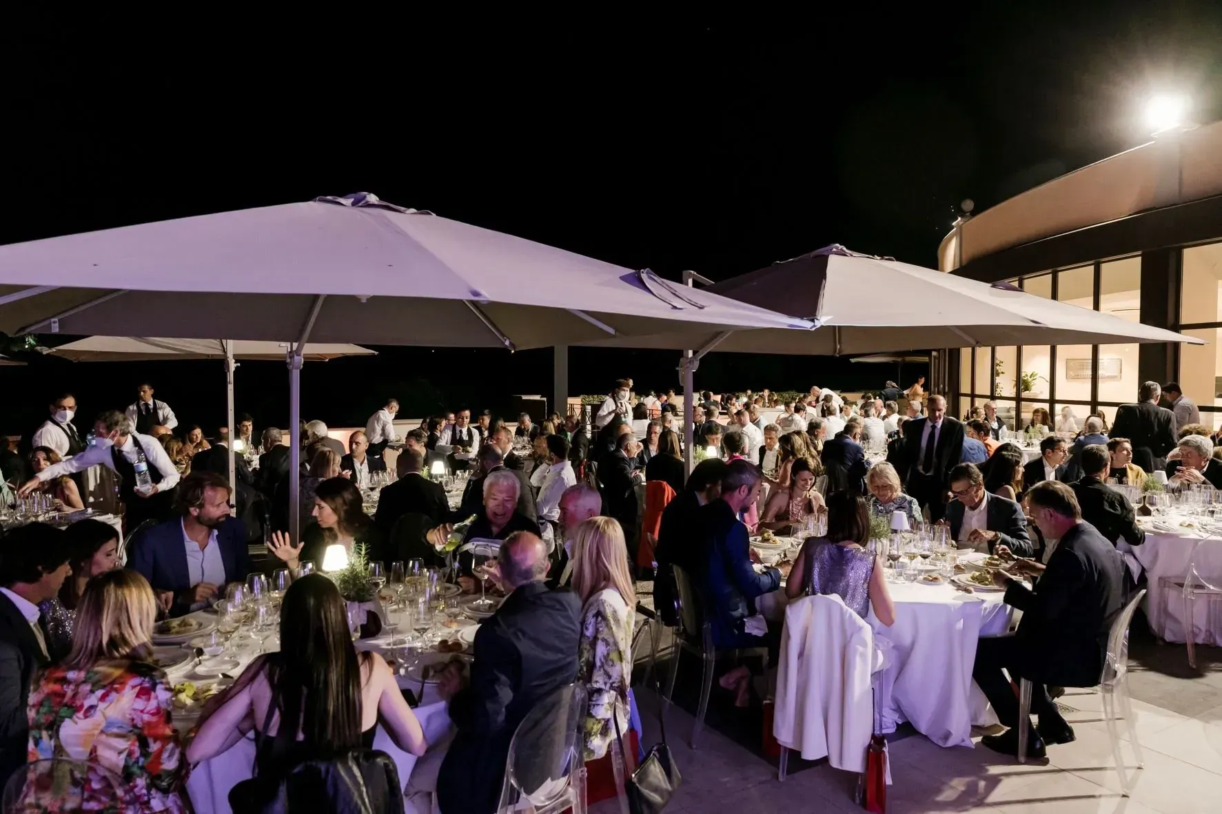 Evento serale all'aperto con molte persone sedute ai tavoli, sotto gli ombrelloni, vicino a un edificio.